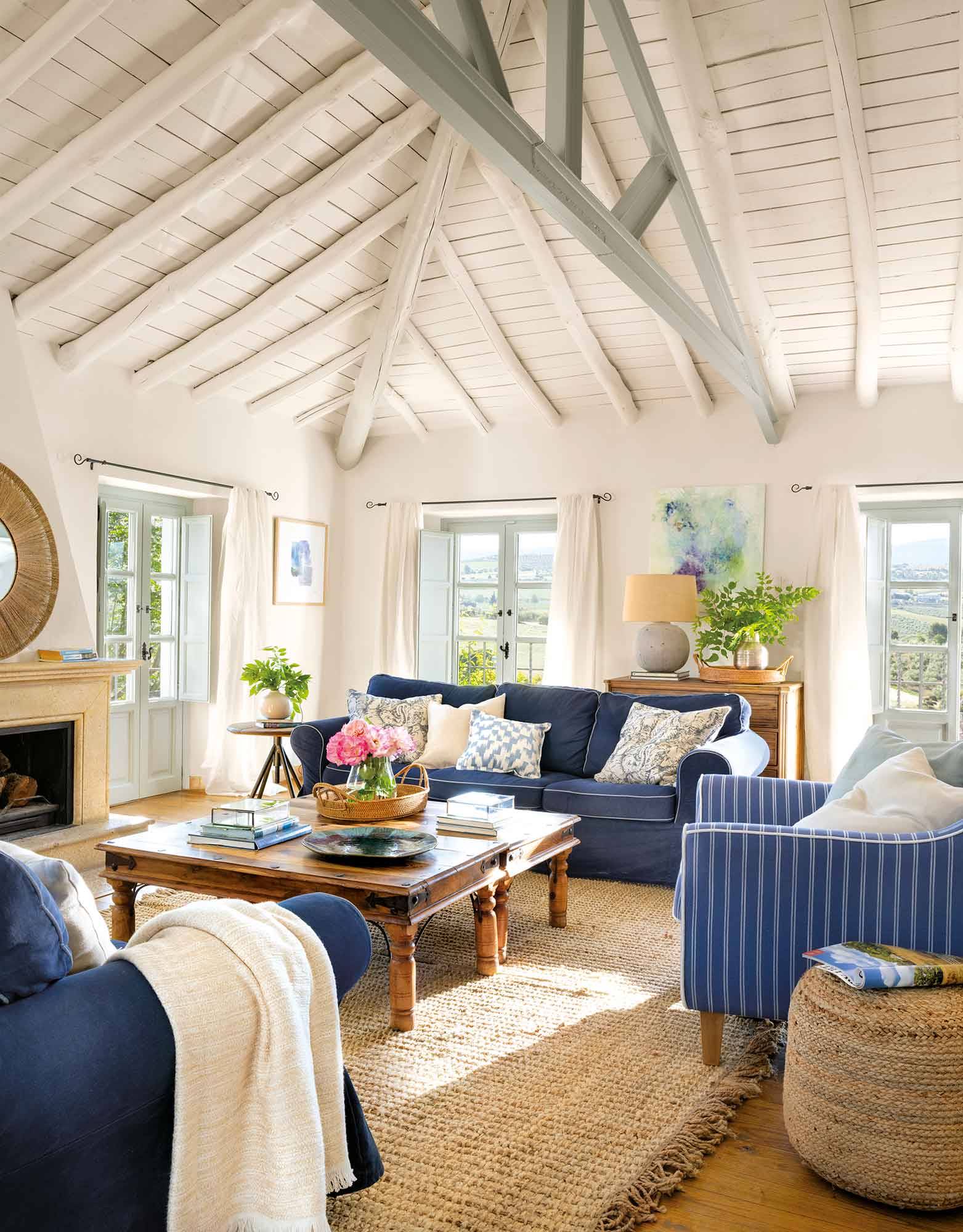 Salón con sofás azules, mesa de madera y techo abuhardillado de color blanco. 