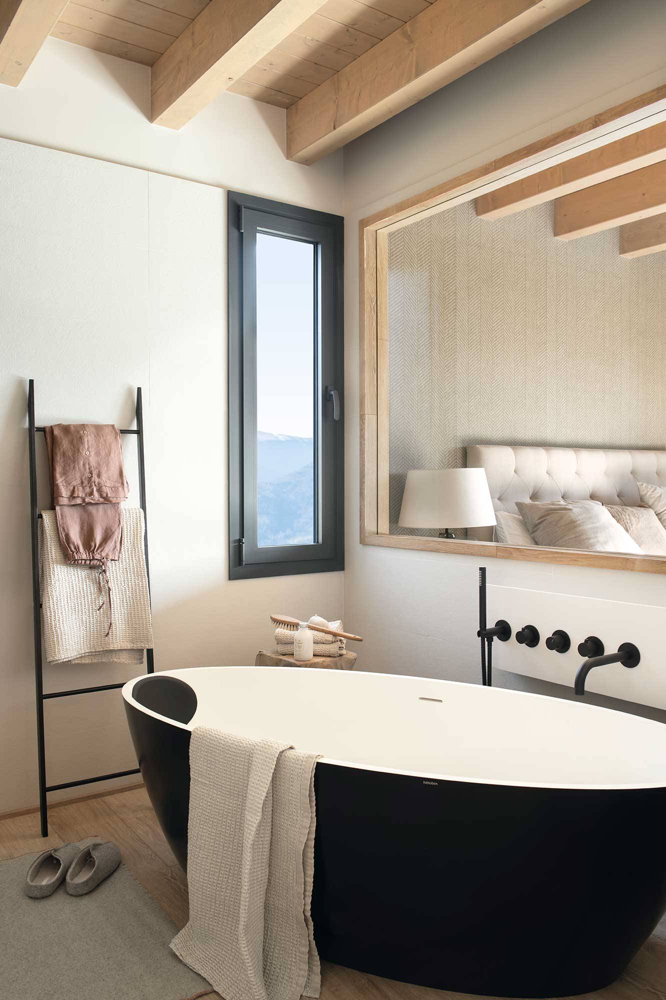 Baño rústico moderno con bañera exenta negra y ventana fija que comunica el baño con el dormitorio. 