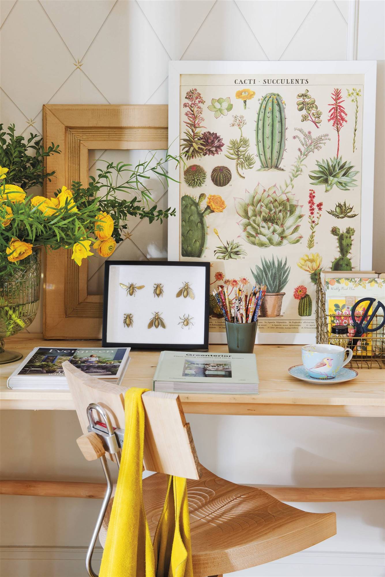 Zona de trabajo con escritorio de madera, silla, ramo de flores y cuadros de cactus e insectos. 