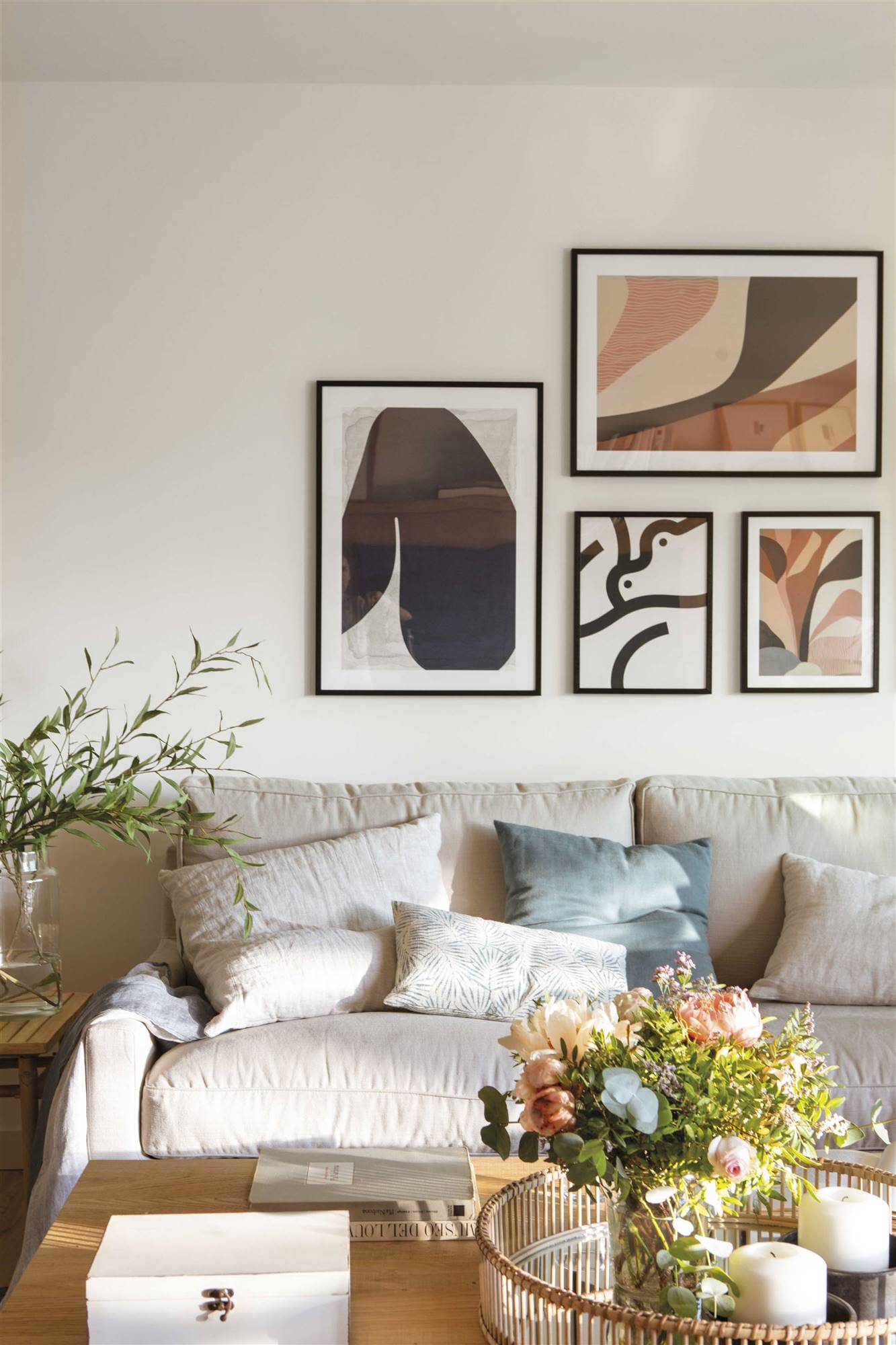 Detalle de salón con sofá beige, cojines , mesa de centro con flores e ilustraciones enmarcadas en la pared. 