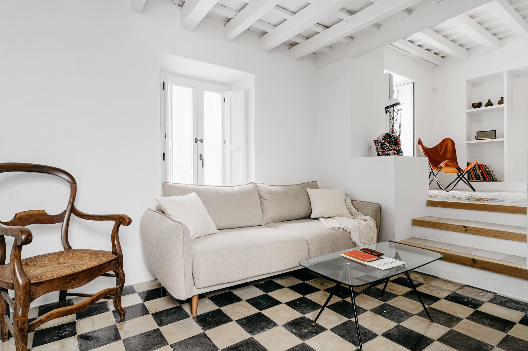 Salón después de la reforma con suelo hidráulico original, sofá beige, silla de madera antigua y paredes blancas.