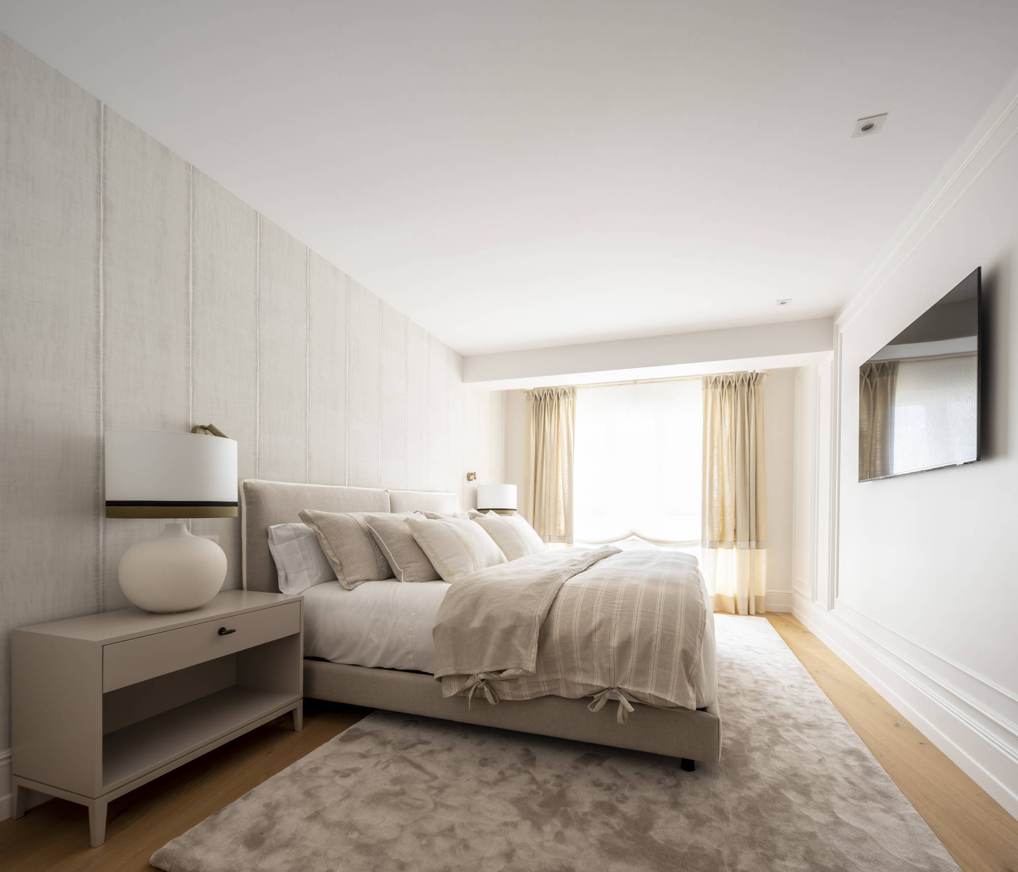 Dormitorio principal en tonos claros con alfombra, papel pintado y televisor.