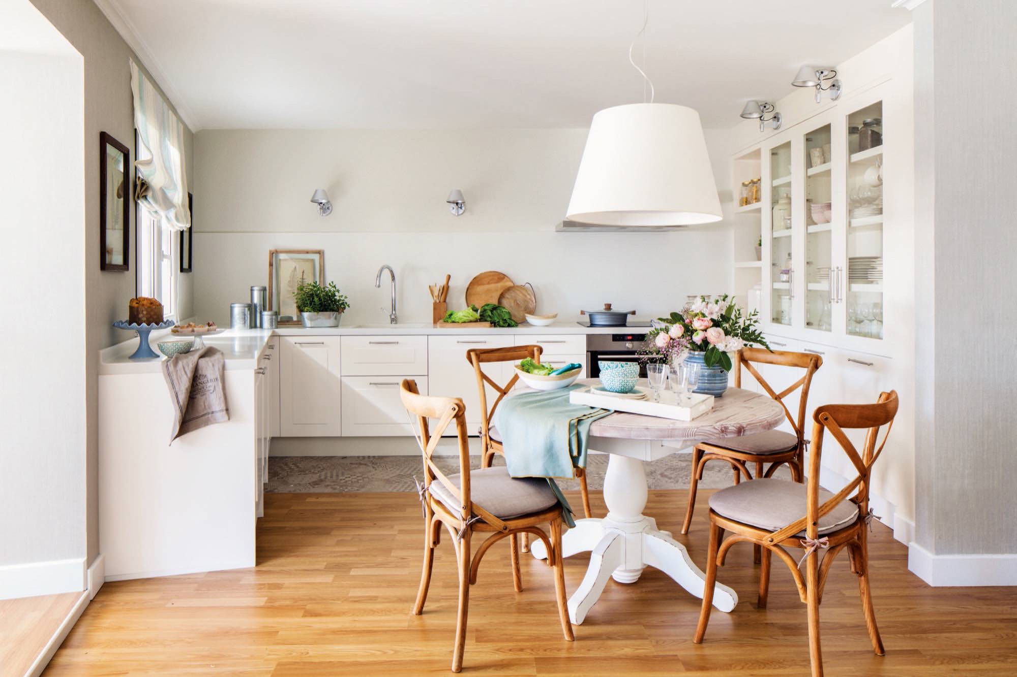 Cocina blanca con vitrina y office con mesa de comedor redonda antigua y sillas de madera.