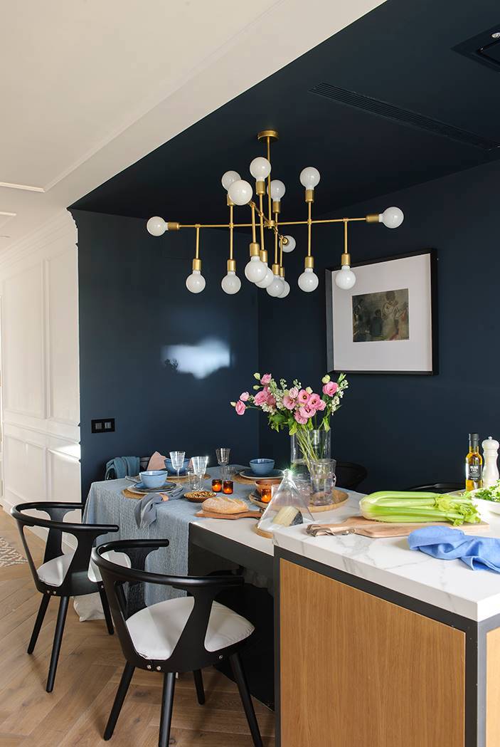 Comedor abierto a la cocina con pared azul intenso, lámpara dorada y sillas negras 00470695
