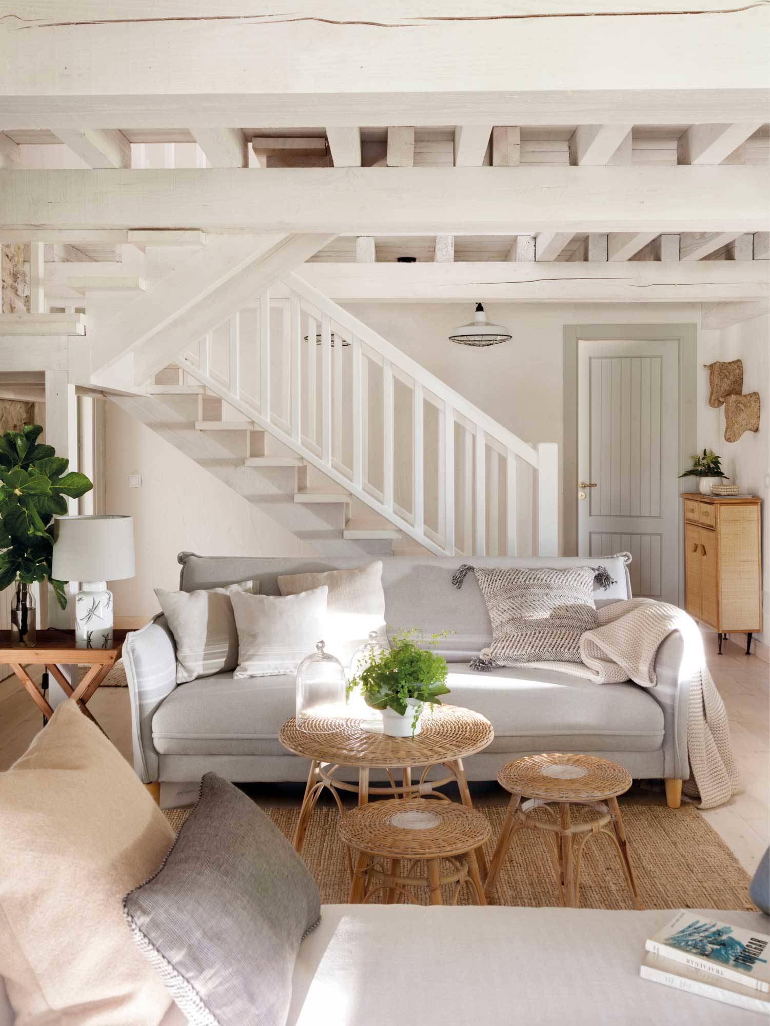 Salón con sofá gris, mesas nido de fibra y escalera al fondo.