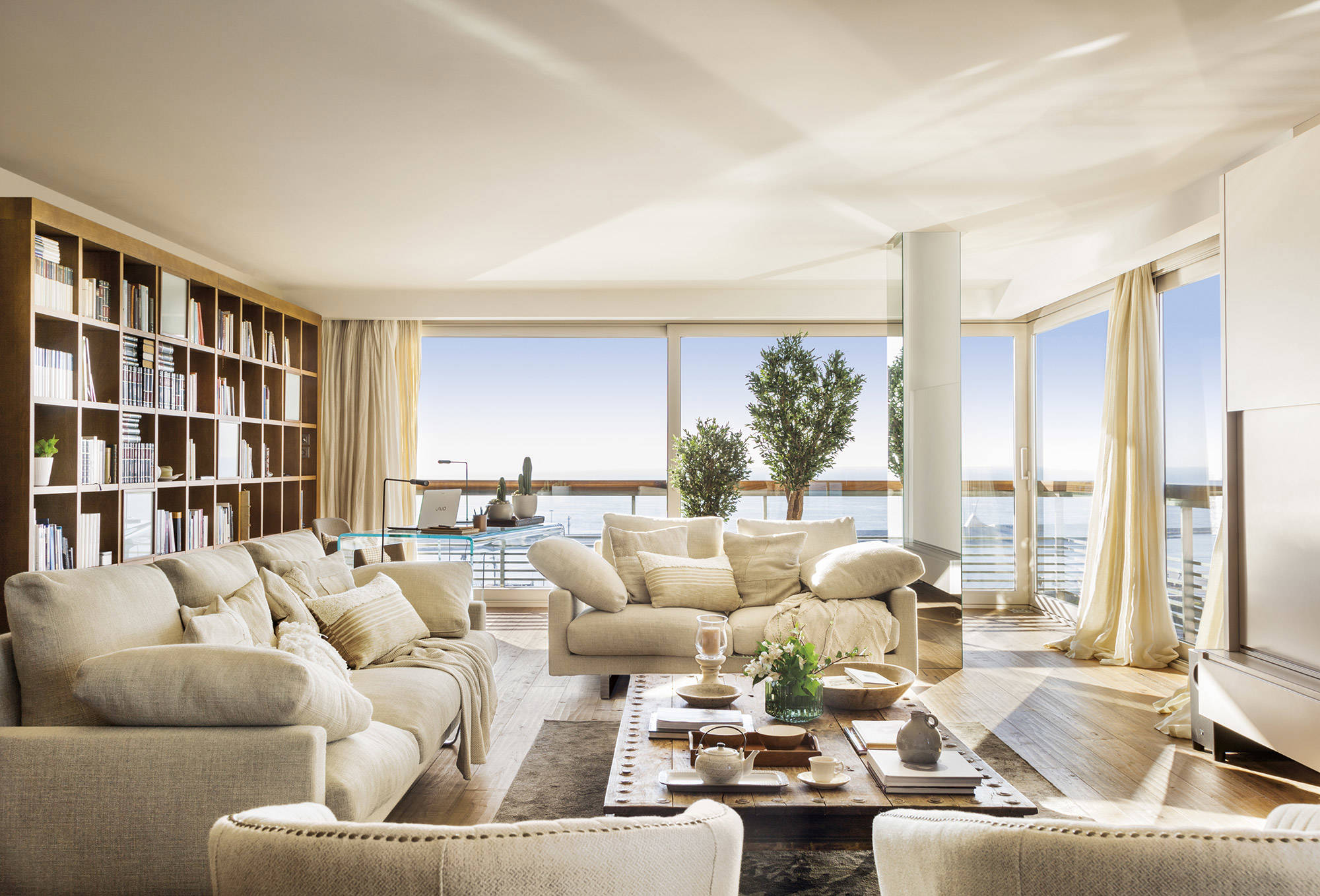 Salón con vistas al mar y grandes ventanales con cortinas beige hasta el suelo. 