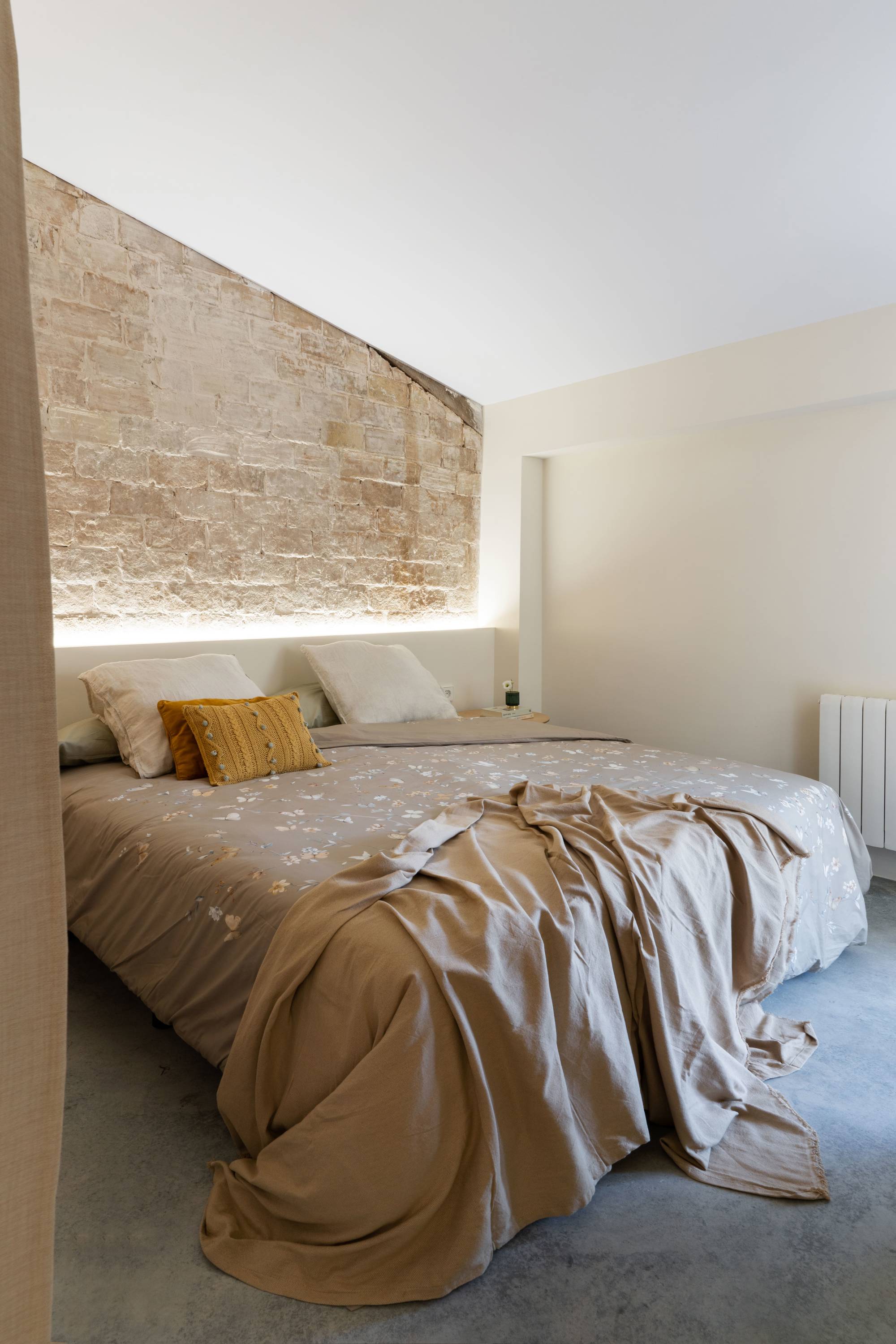 Dormitorio después de la reforma con pared de ladrillos, cabecero iluminado y cama XXL con cojines mostaza y plaid.