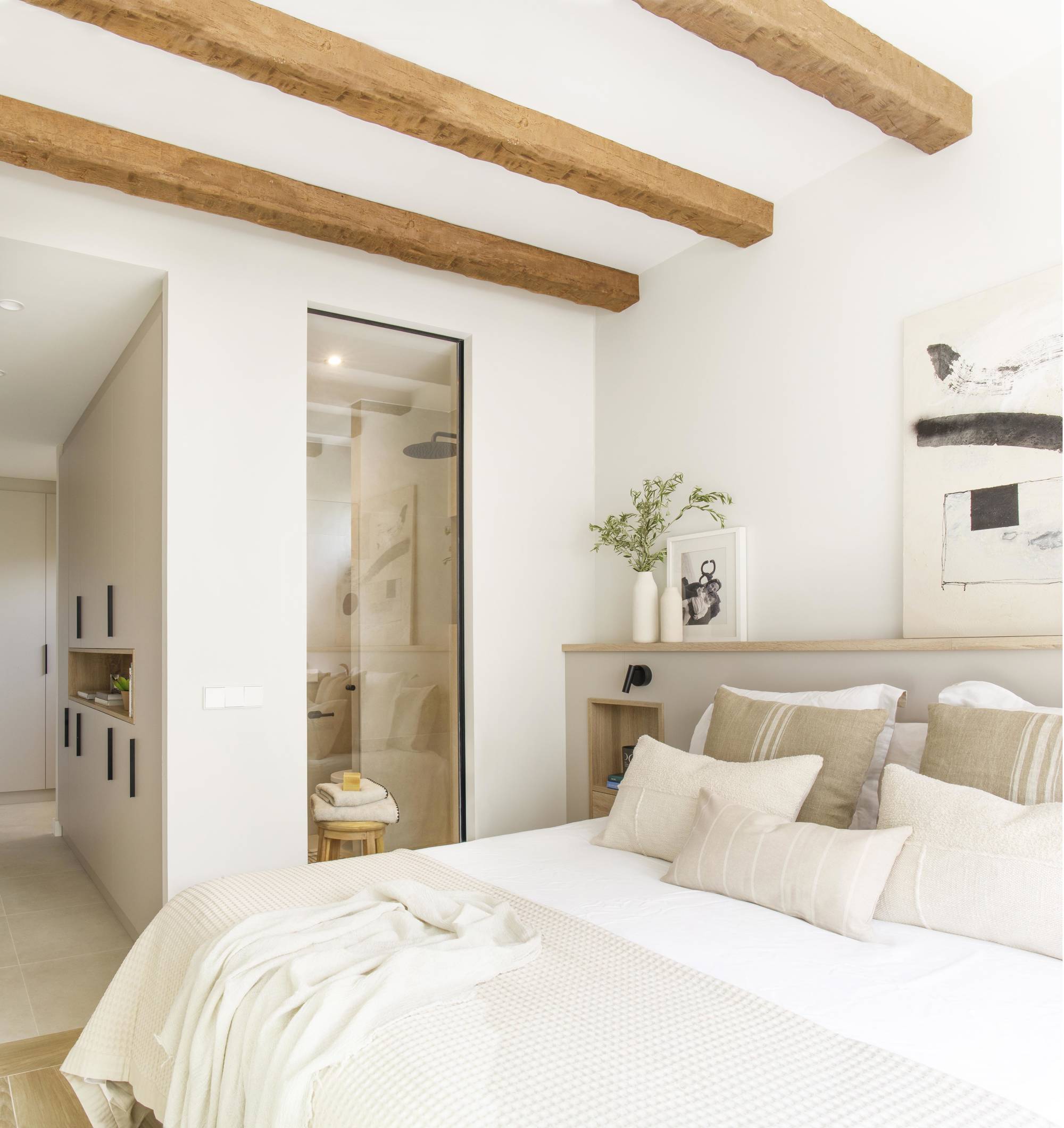 Dormitorio principal en tonos neutros con vigas en el techo, cabecero con hornacinas y cristalera que da al baño. 
