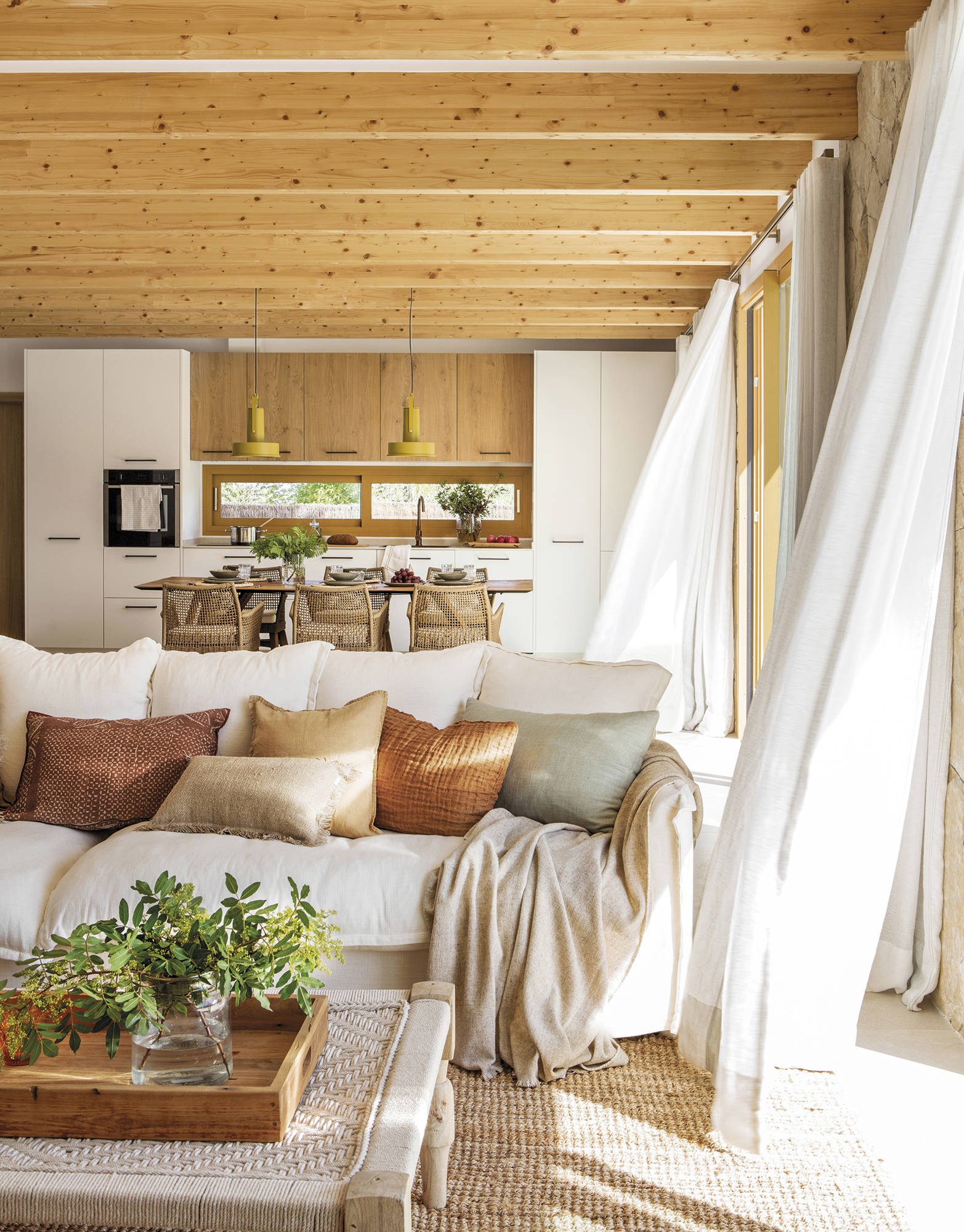 Salón abierto a la cocina con sofá blanco y techo de vigas de madera.