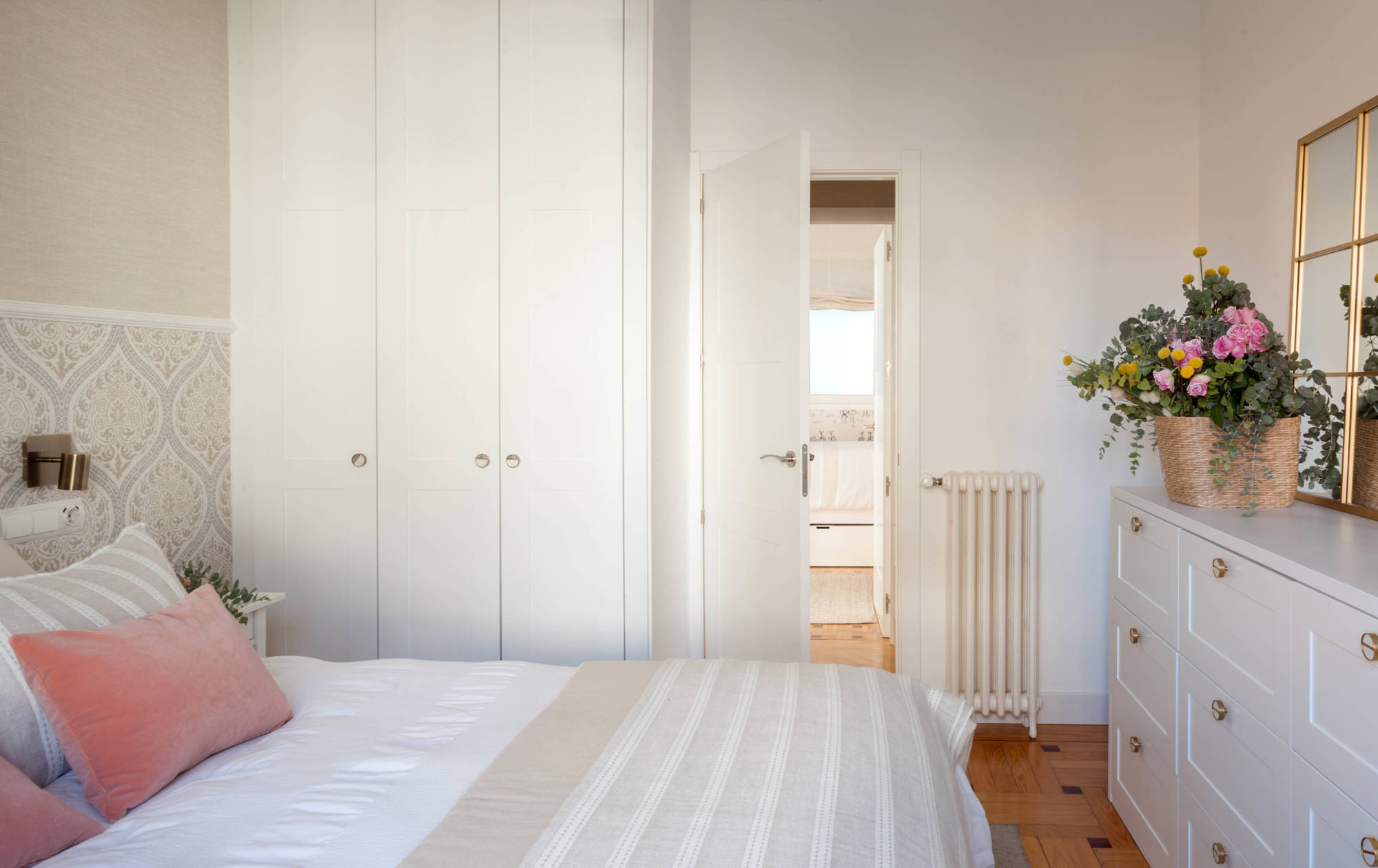 Dormitorio principal con armarios, cómoda, flores, cojines rosas empolvados y papel pintado.