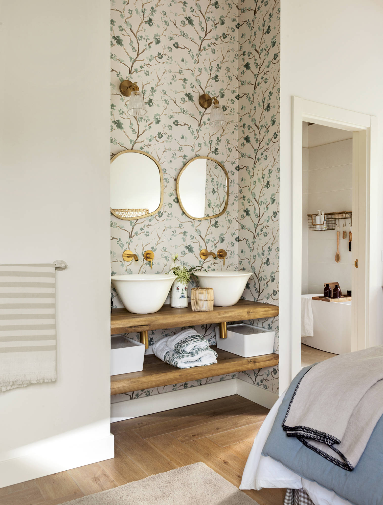 Dormitorio con baño en la habitación con dos lavamanos, dos espejos, baldas de madera abiertas y papel pintado. 