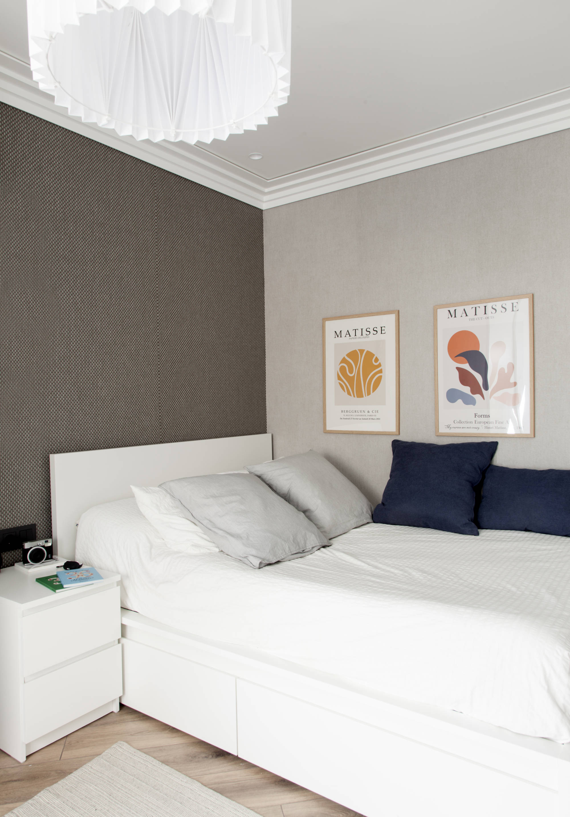 Habitación juvenil con cama grande, cuadros, cojines azules y pared del cabecero gris oscuro con papel pintado.