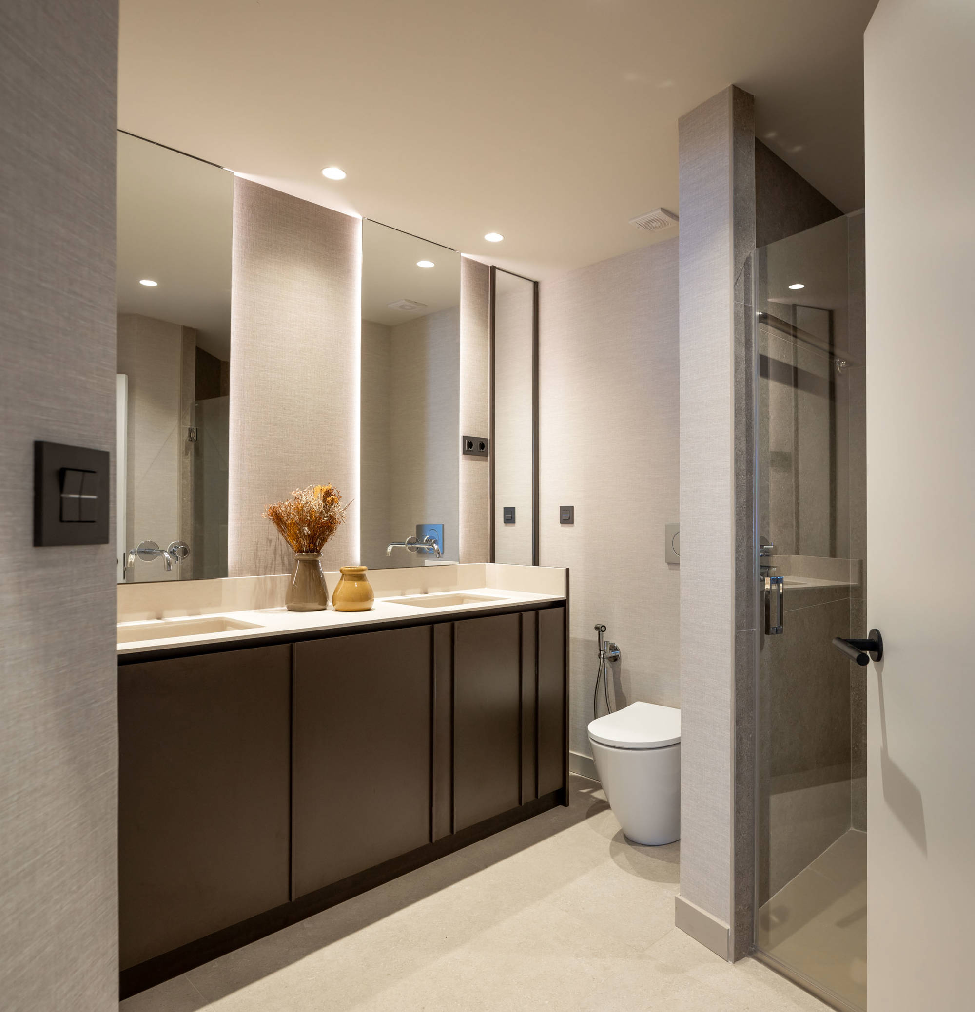 Baño con mueble bajolavabo con doble lavabo de madera, espejos iluminados y zona de ducha con mampara. 