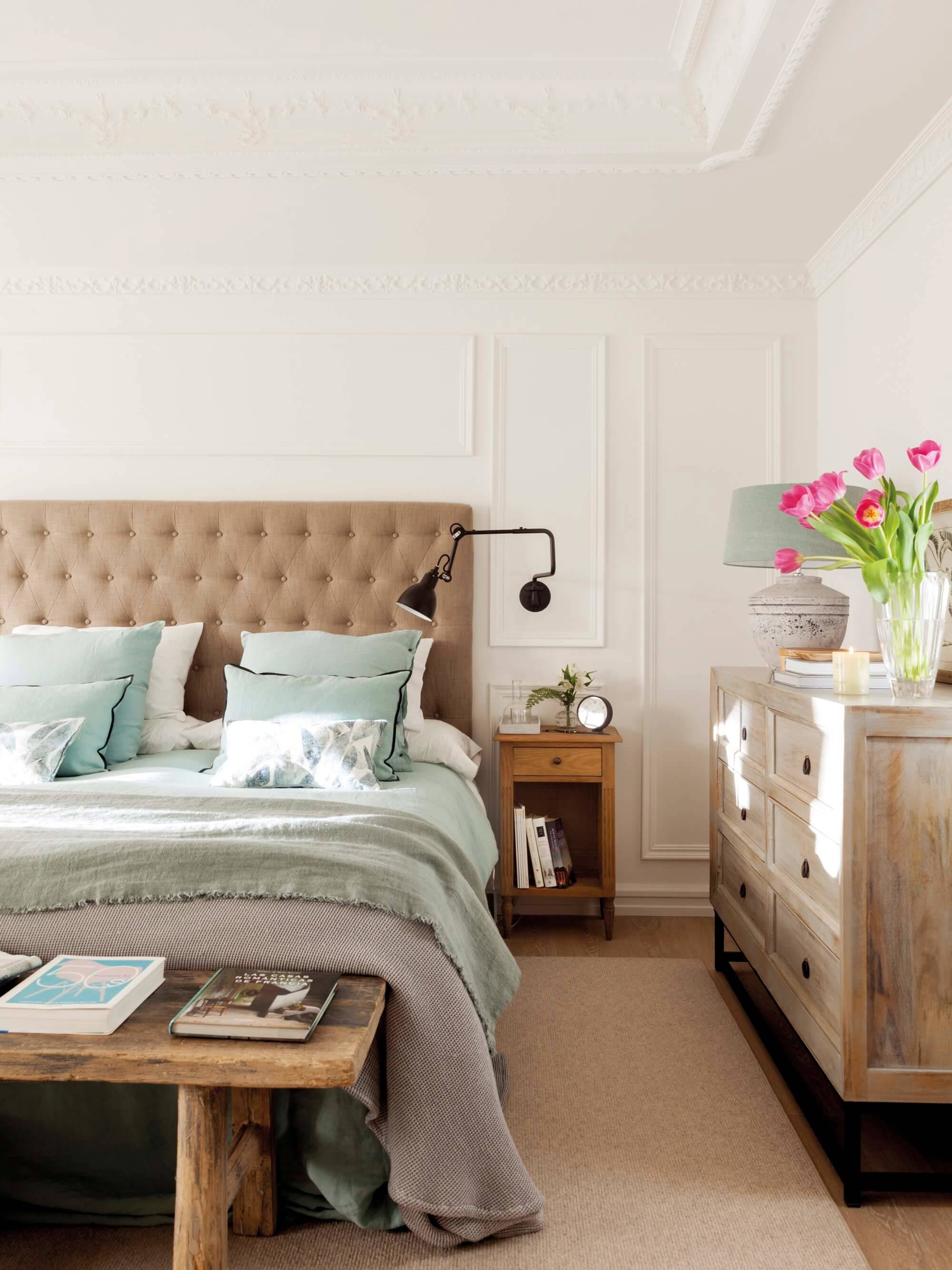 Dormitorio con cabecero con tapizado capitoné, banco de madera, cómoda de madera decapada y molduras decorativas.