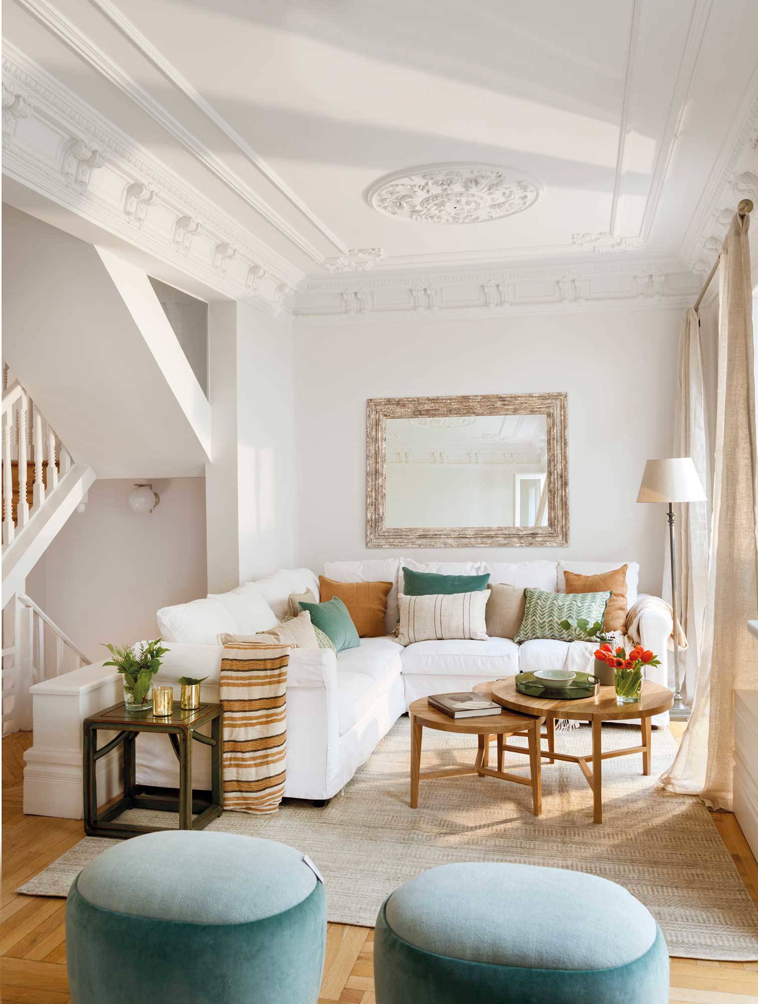 Salón con sofá rinconera blanco, mesas nido de madera y puffs de terciopelo azul.