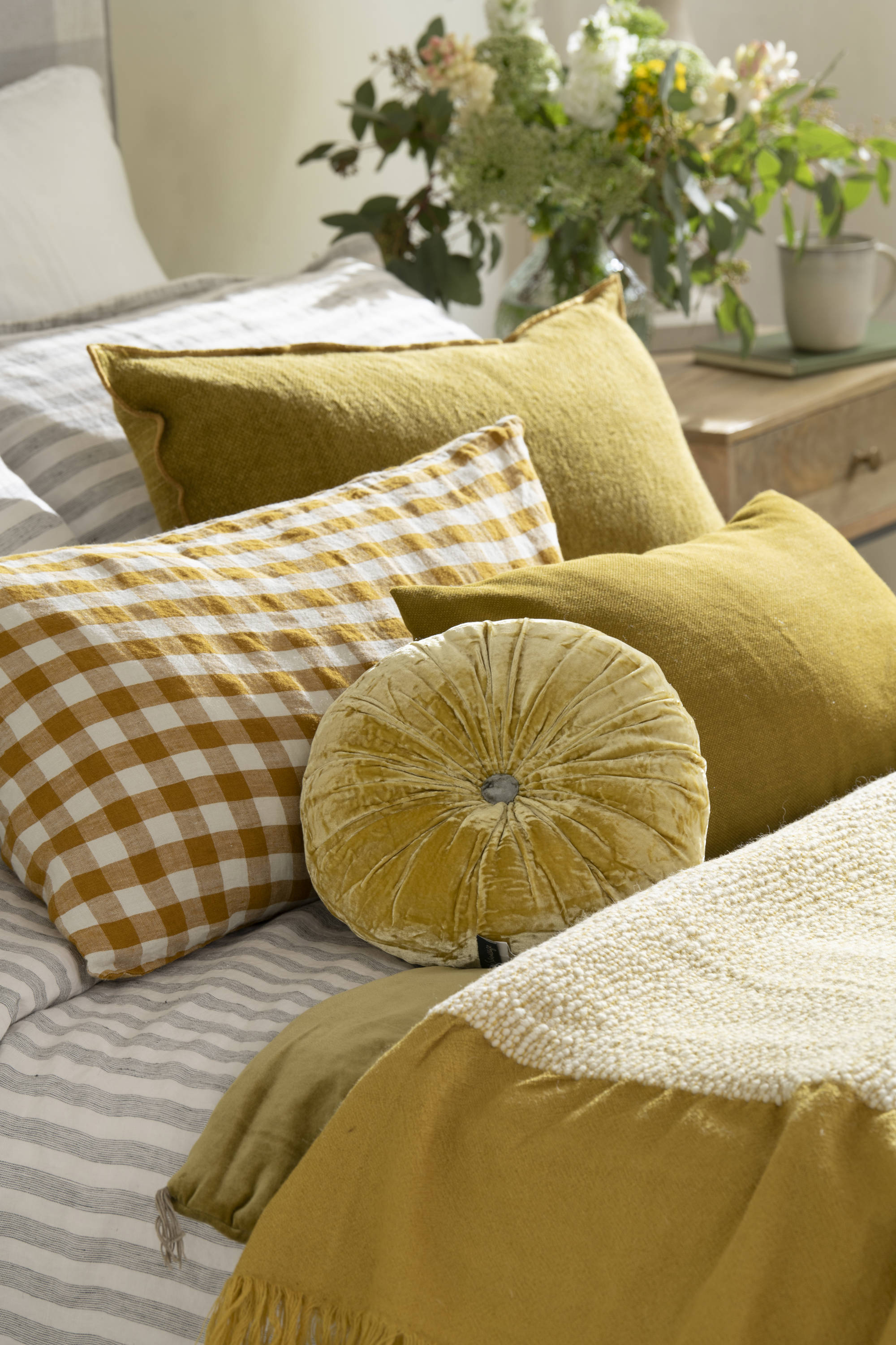 Dormitorio con cama con cojines mostaza, funda nórdica a rayas en gris y jarrón de flores. 