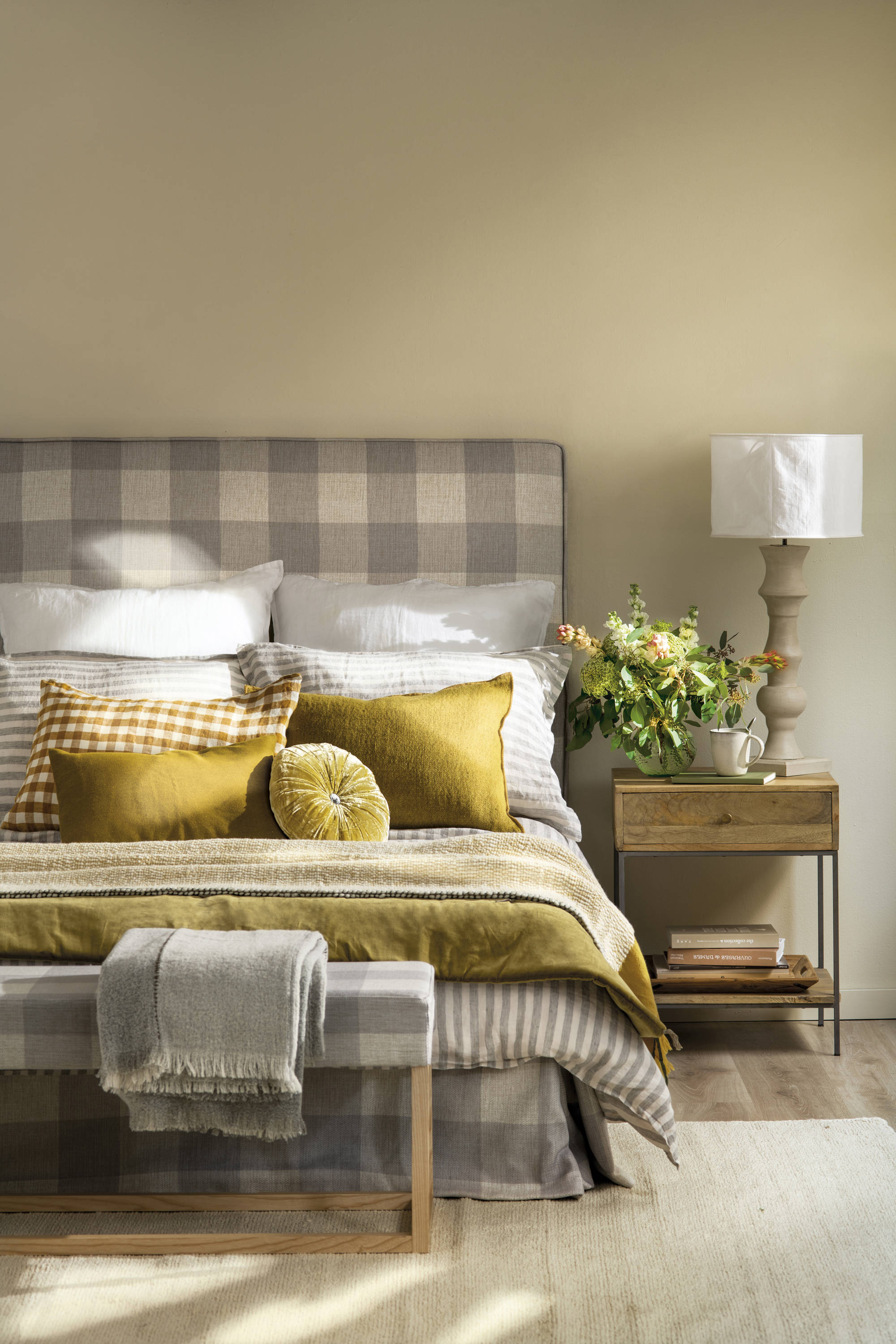 Dormitorio con cama con ropa de cama mostaza, cabecero tapizado a cuadros y mesita de noche de madera. 