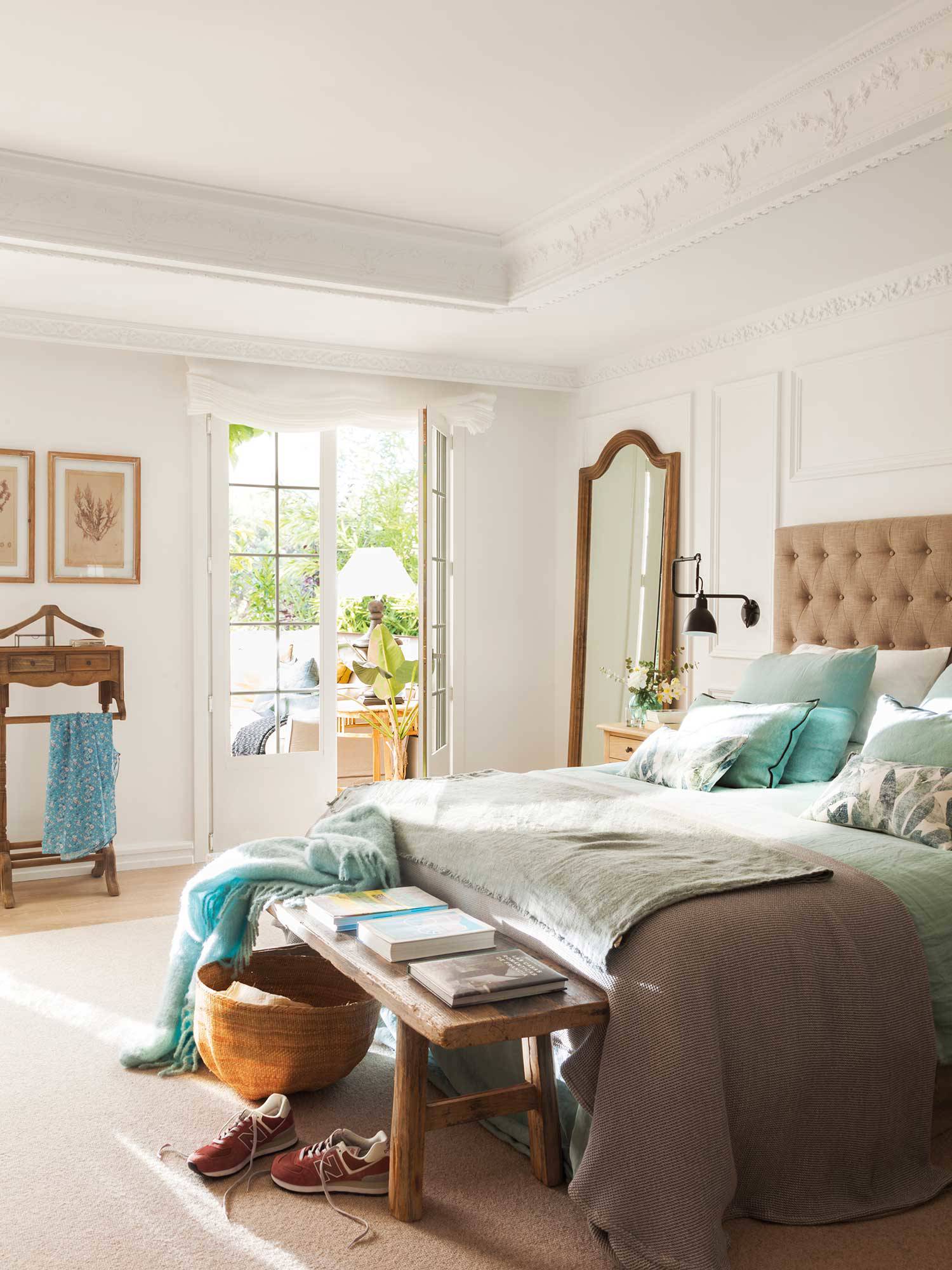 Dormitorio clásico actualizado con cabecero capitoné, banco de madera, galán de noche y espejo con marco de madera.