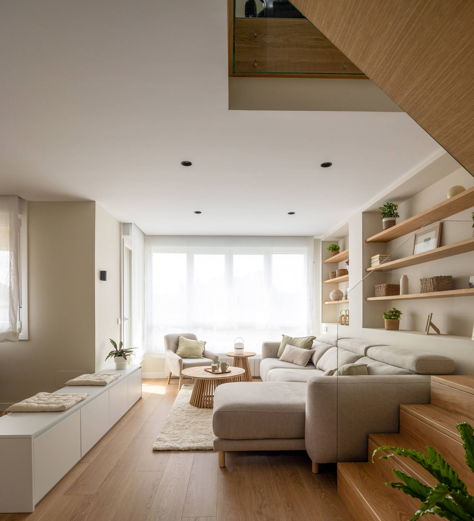 Salón moderno con escalera de madera, sofá beige con chaise longe y ventanal.