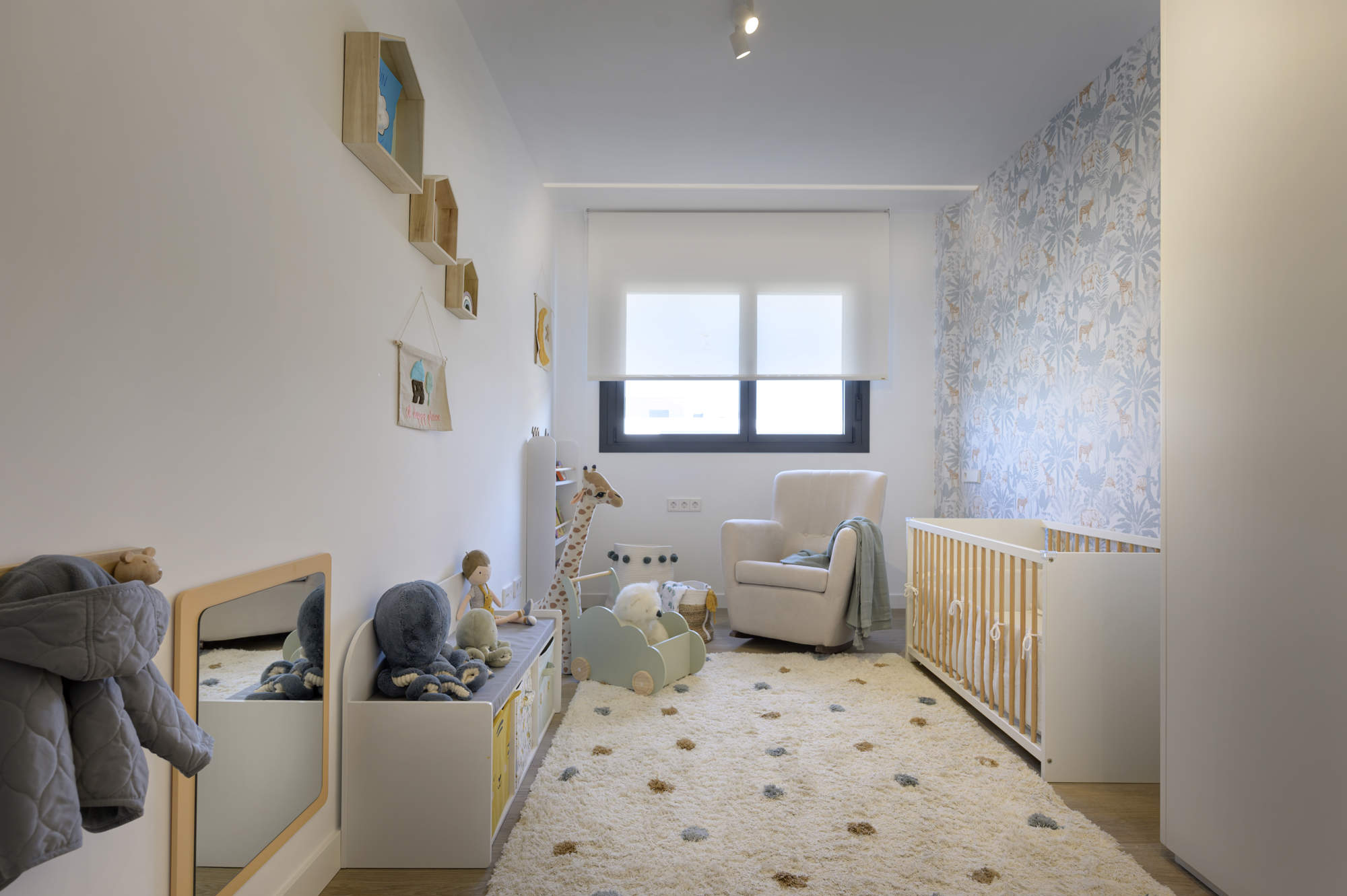 Dormitorio del bebé con alfombra de topos, papel pintado, cuna de madera, butaca gris y muebles Montessori.