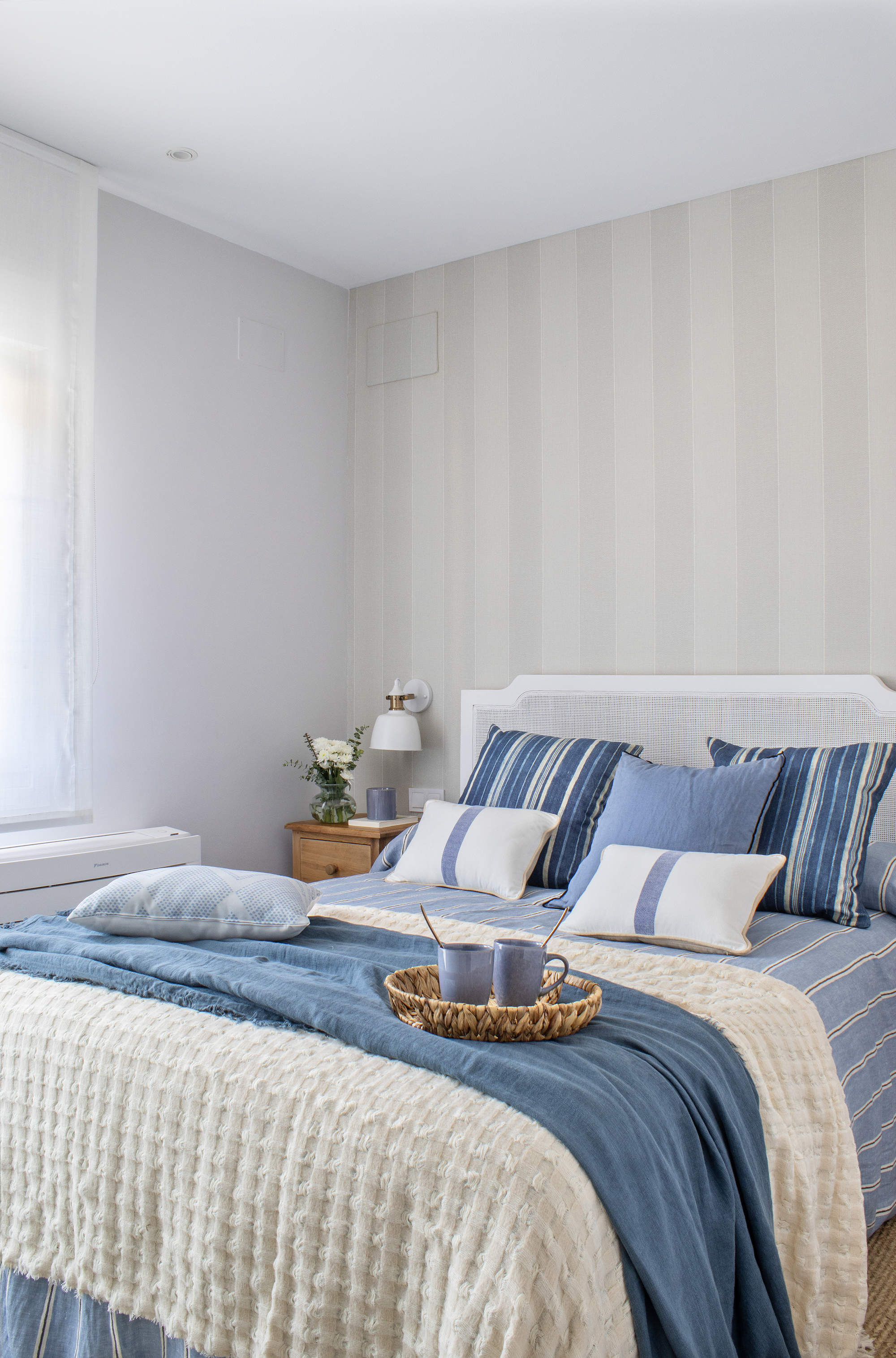 Un dormitorio principal con papel pintado de rayas en color neutro y colcha y cojines en azul y rayas.