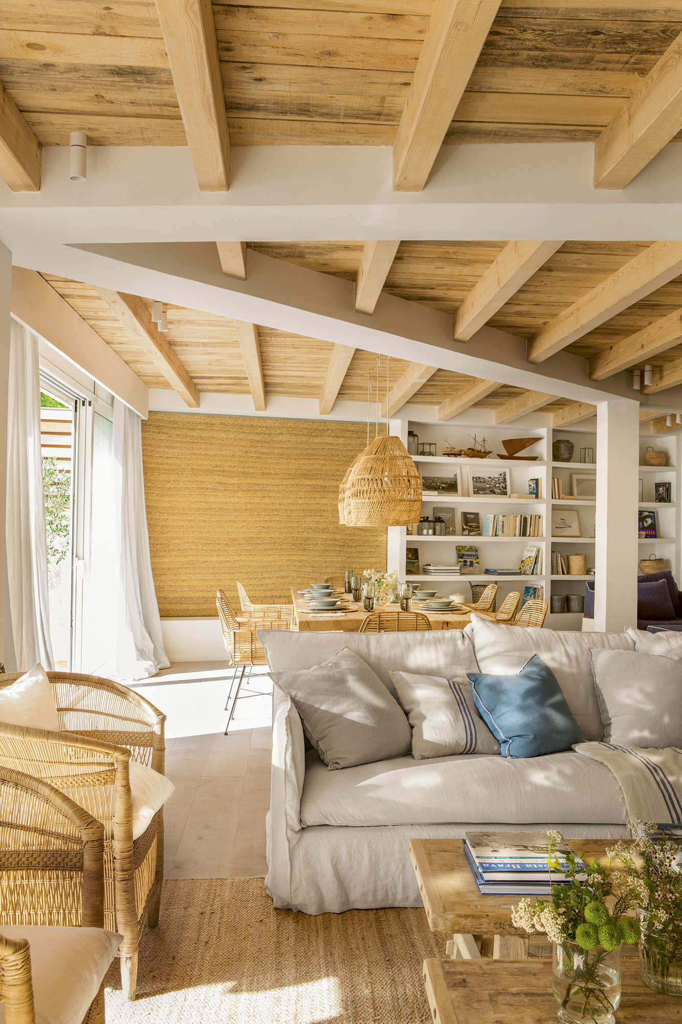 Salón con techo de madera, muebles de fibras y sofá gris con cojines. 