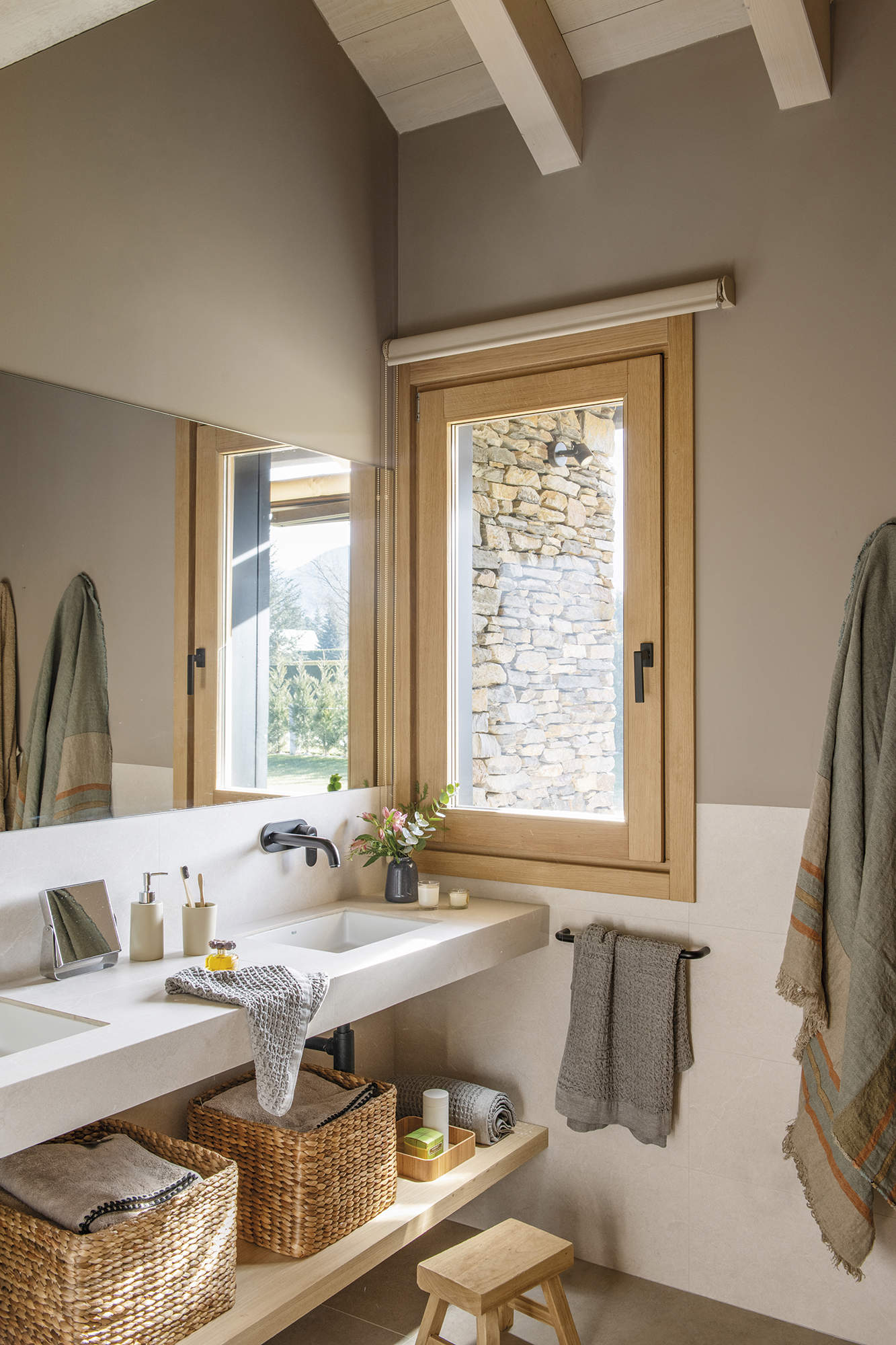 baño con dos lavabos, canasta de fibra con toallas, flores, ventana de madera, pared gris, suelo de gres gris