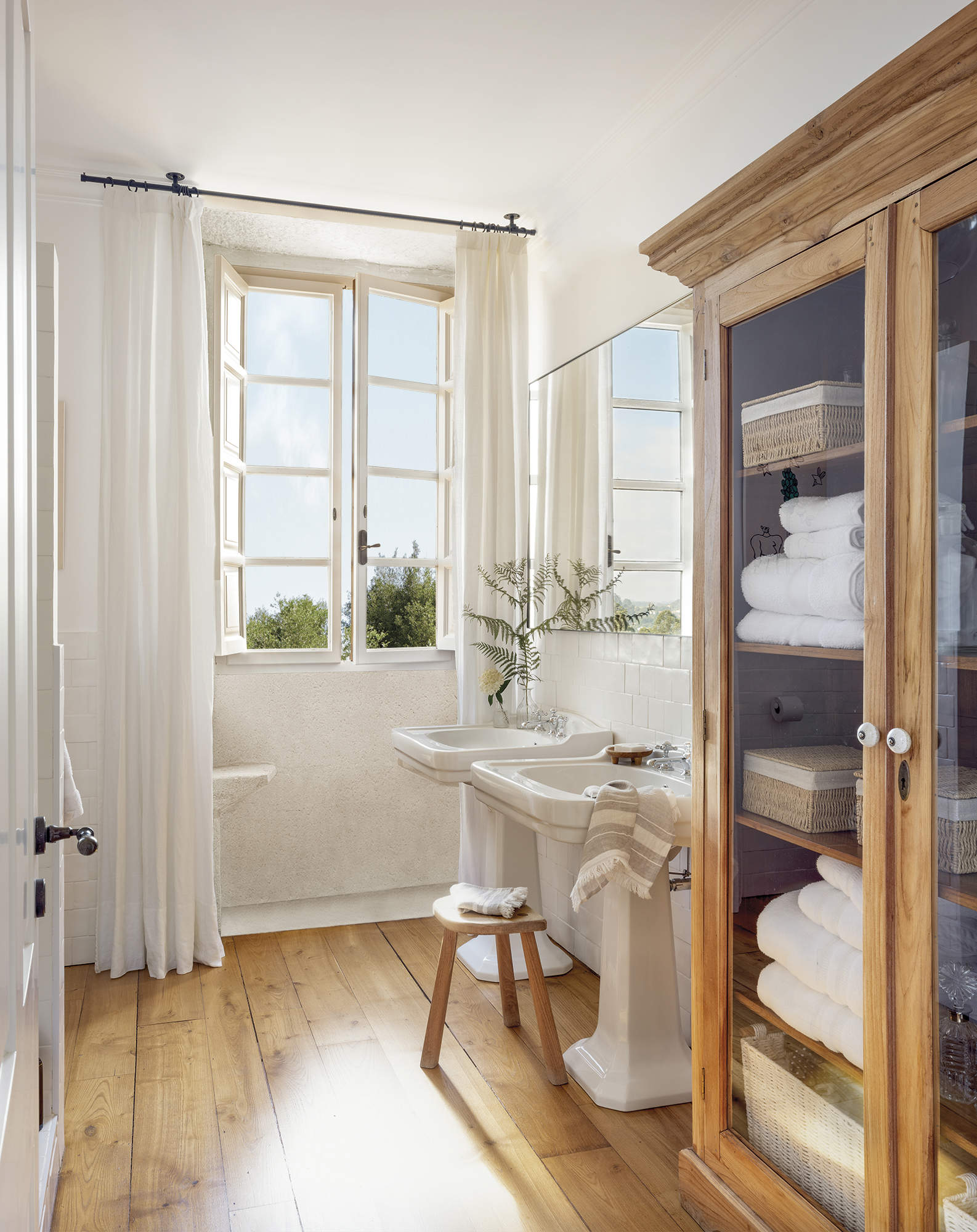 baño con suelo de madera y dos lavabos, cortinas blancas, vitrina con ropa blanca