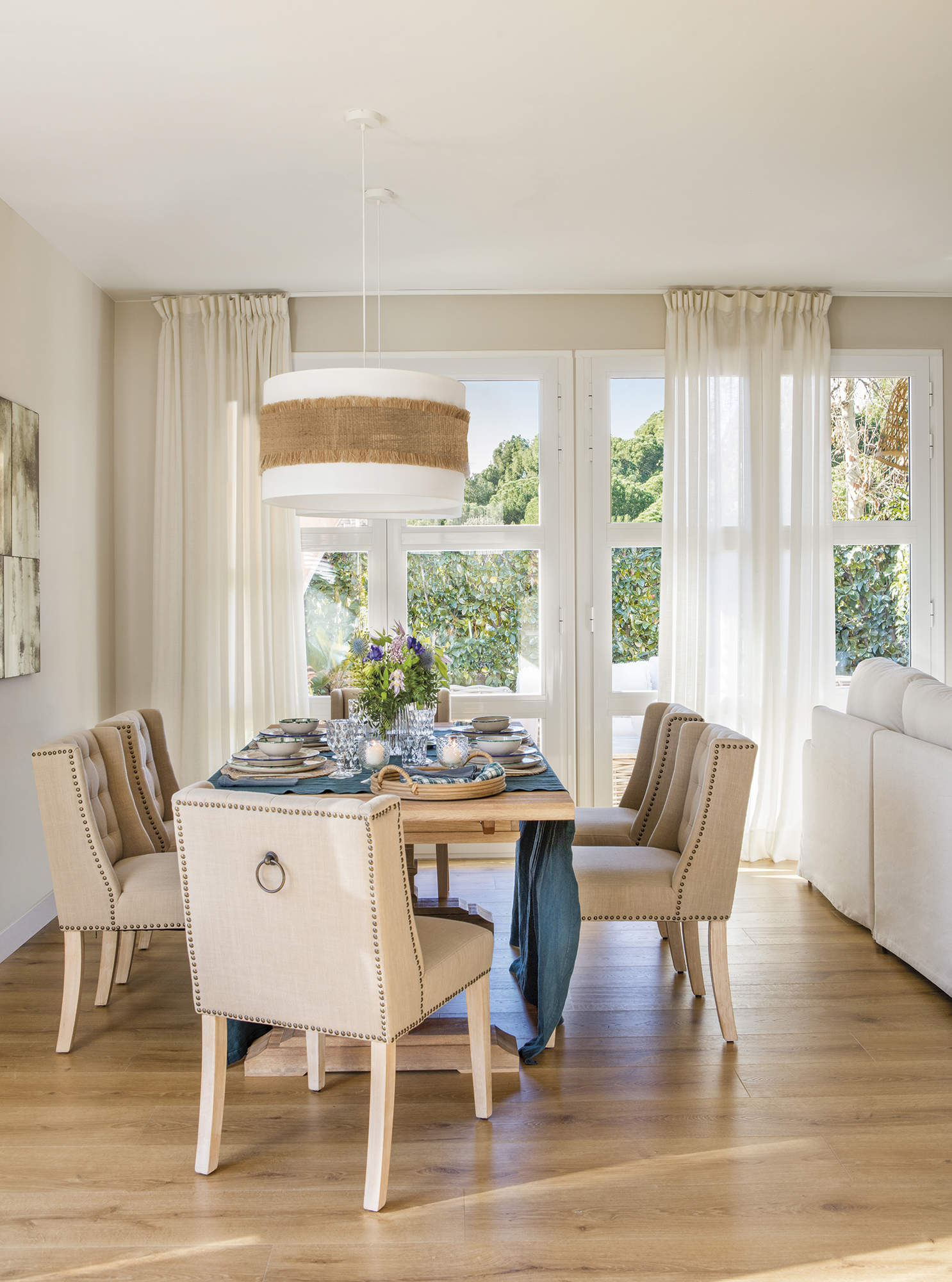 Comedor con mesa rectangular y sillas tapizadas beige, lámpara de techo, salida al jardín con cortinas blancas.