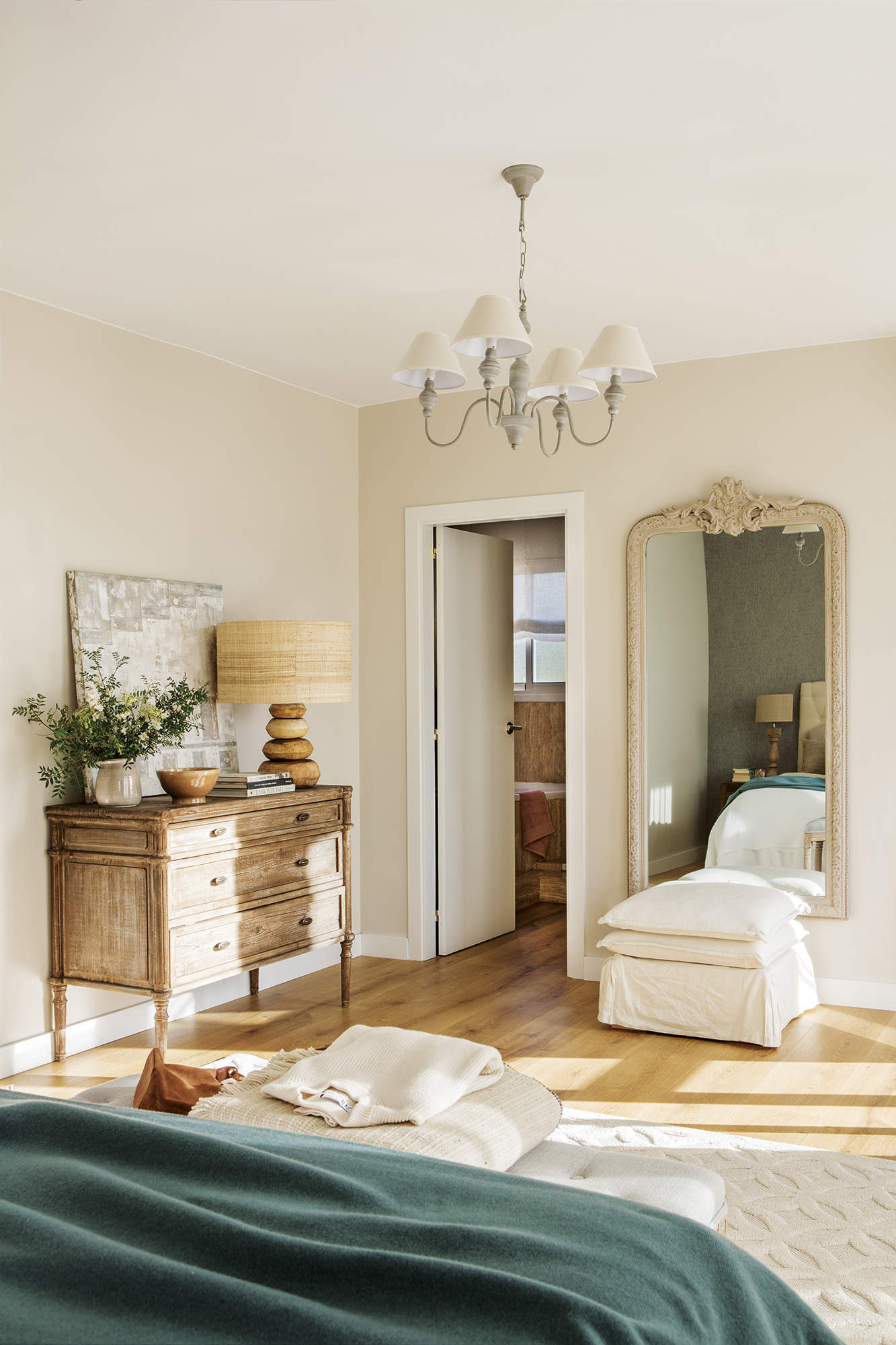 Dormitorio principal con baño, espejo grande, cómoda de madera envejecida, lámpara de mesa de madera, lámpara de techo clásica, cuadro, suelo de madera y puf tapizado.