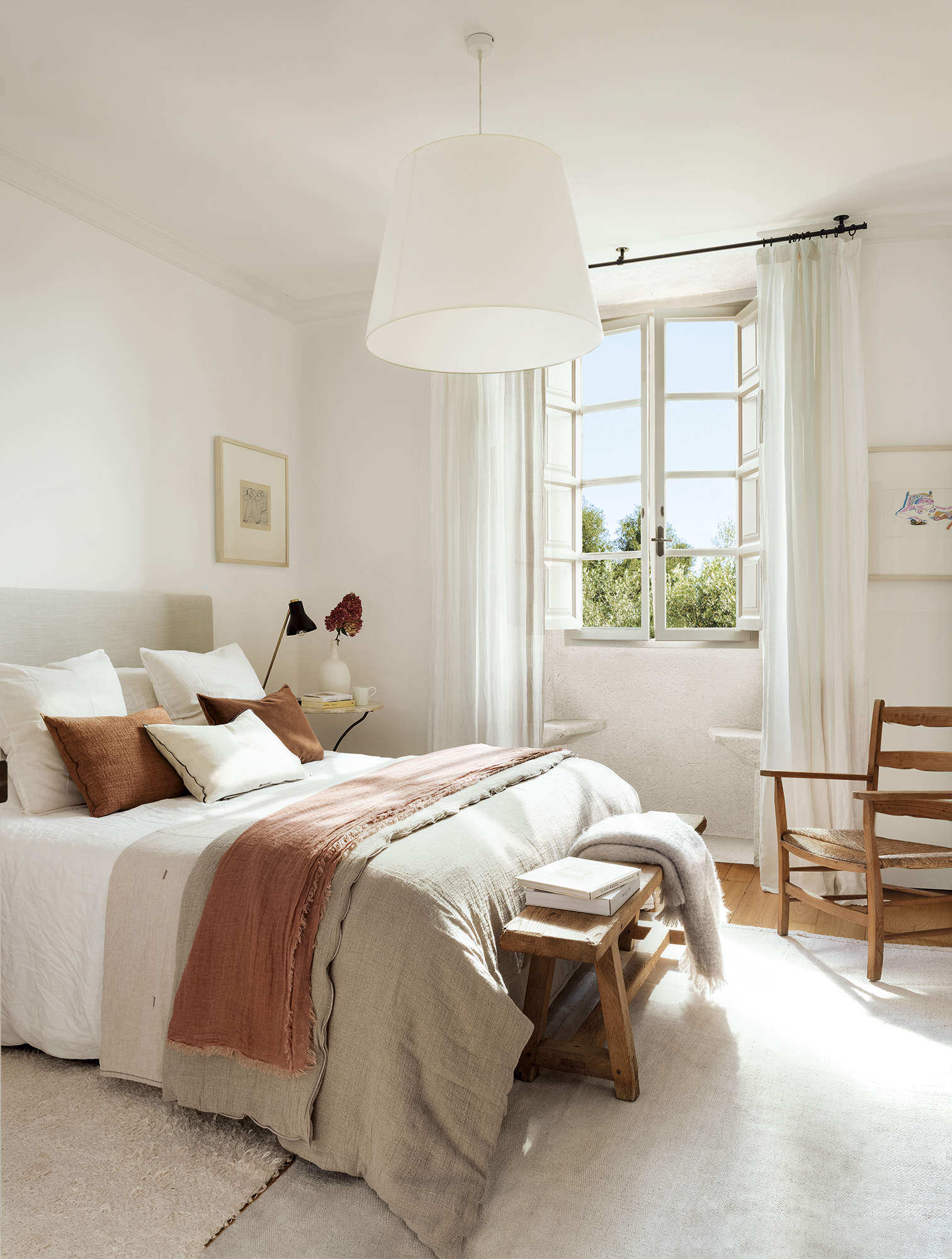 dormitorio principal con ventanal, suelo de madera con alfombra beige, lámpara de techo blanca, ropa de cama beige, cabezal tapizado, banco de madera