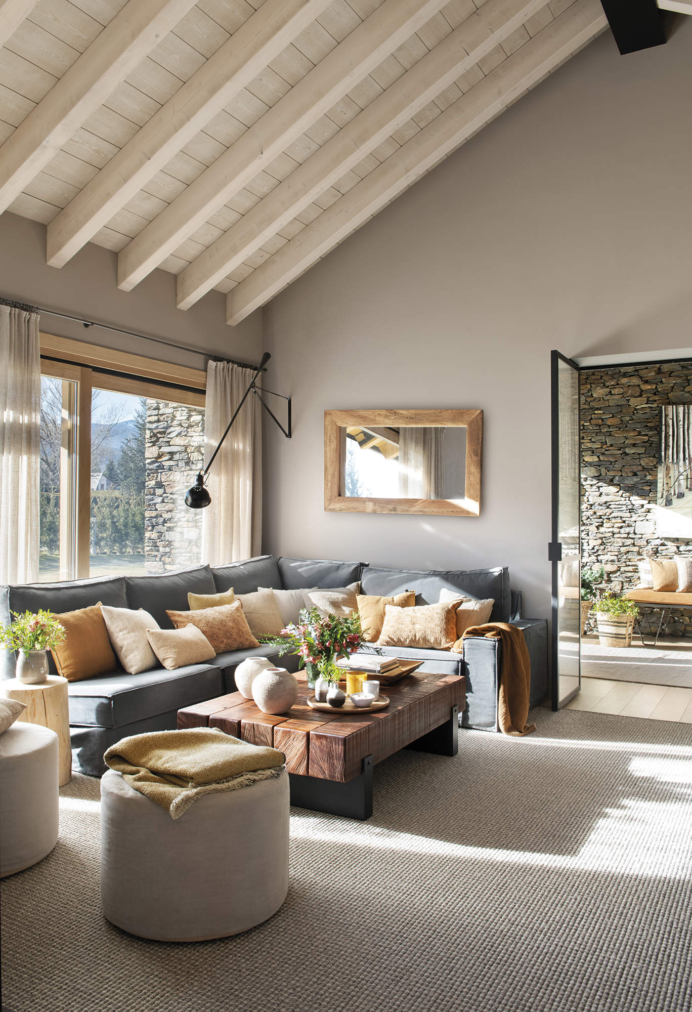 salón con techo alto con vigas de madera, sofá gris esquinero con cojines beige y naranja, mesa de centro de madera rústica,  alfombra gris