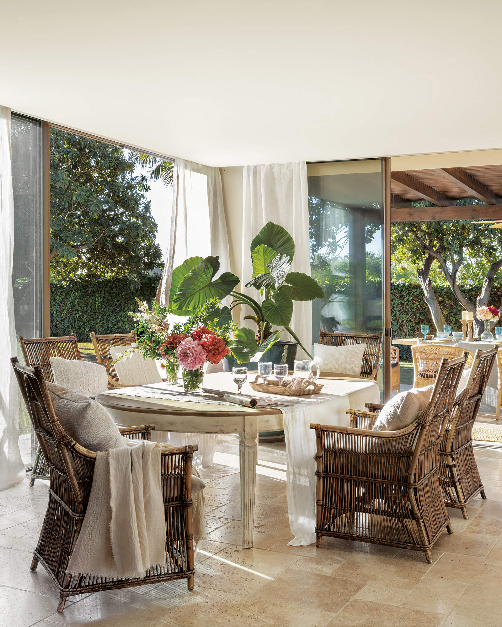 comedor con mesa de madera decapada y sillas de mimbre y bambú, plantas, puerta de cristal con salida al porche, bandeja con copas