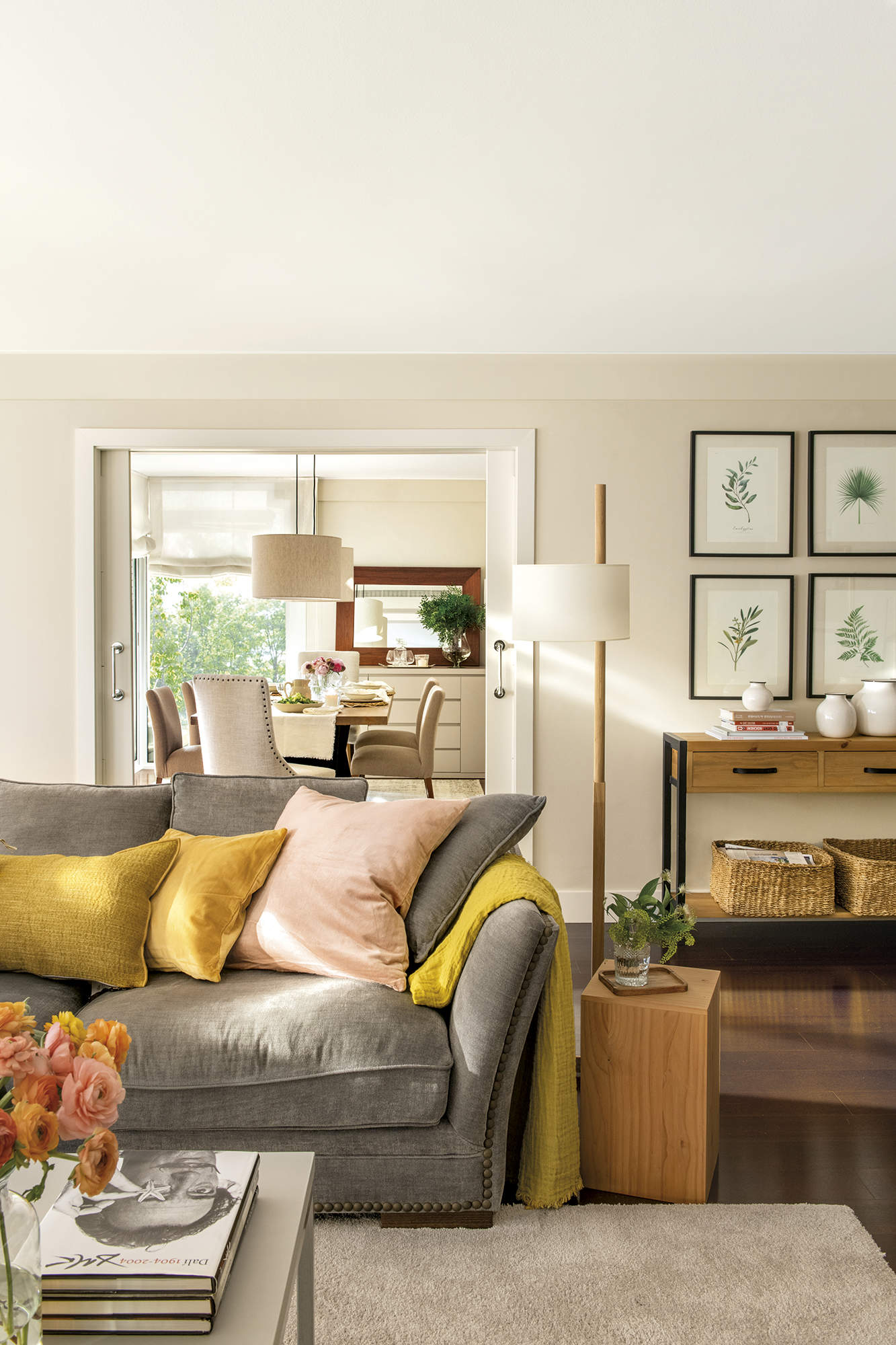 Salón con comedor al fondo, sofá gris con cojines de colores, puerta corredera, lámpara de pié, mueble con cuadros botánicos.