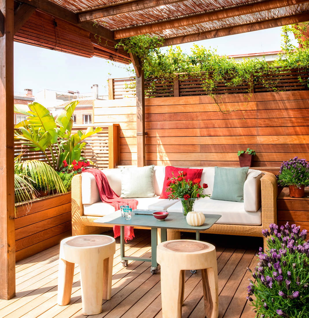 Terraza con tarima, pared de lamas de madera horizontal, sofá de fibra, taburetes inspiración tronco, plantas y pérgola de caña.