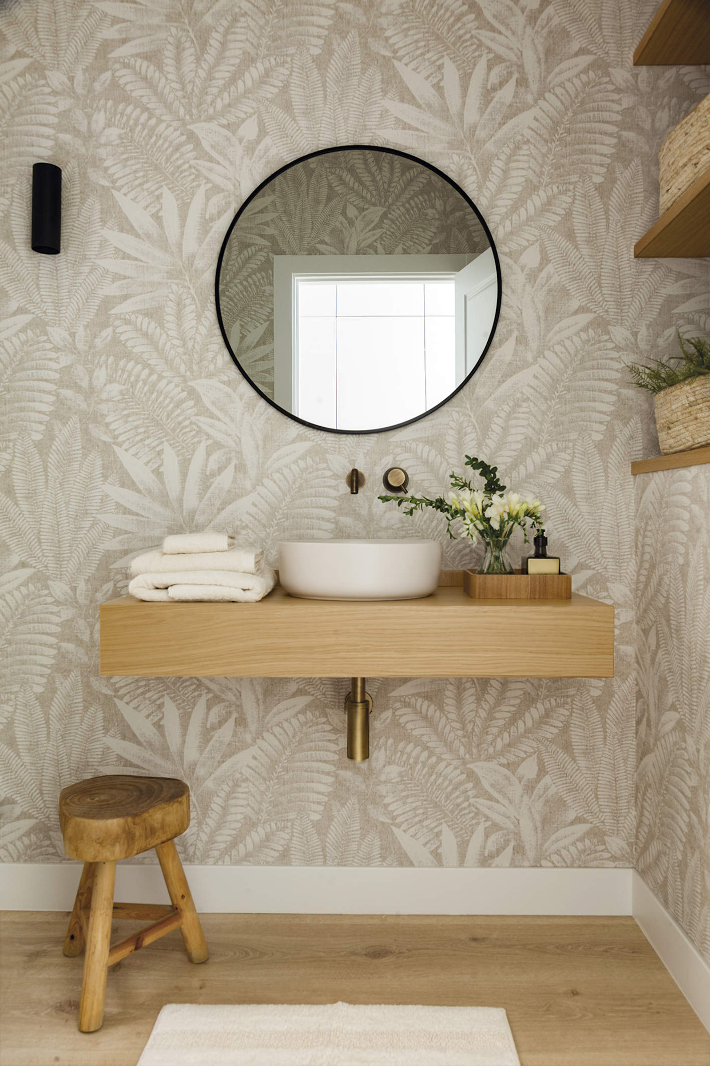 Baño con papel pintado, mueble de madera y taburete. Espejo, redondo. 