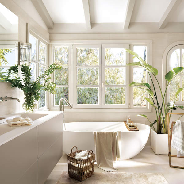 baño grande con bañera externa blanca y mueble de lavabo de dos senos, plantas, suelo de piedra con alfombra beige, gran ventanal