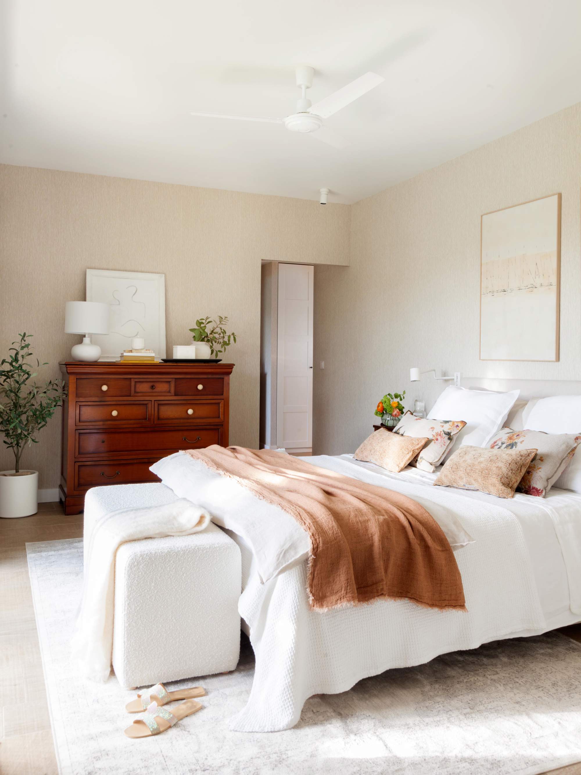 Dormitorio con ropa de cama blanca y rosa, puff a los pies de la cama y cómoda, de madera.