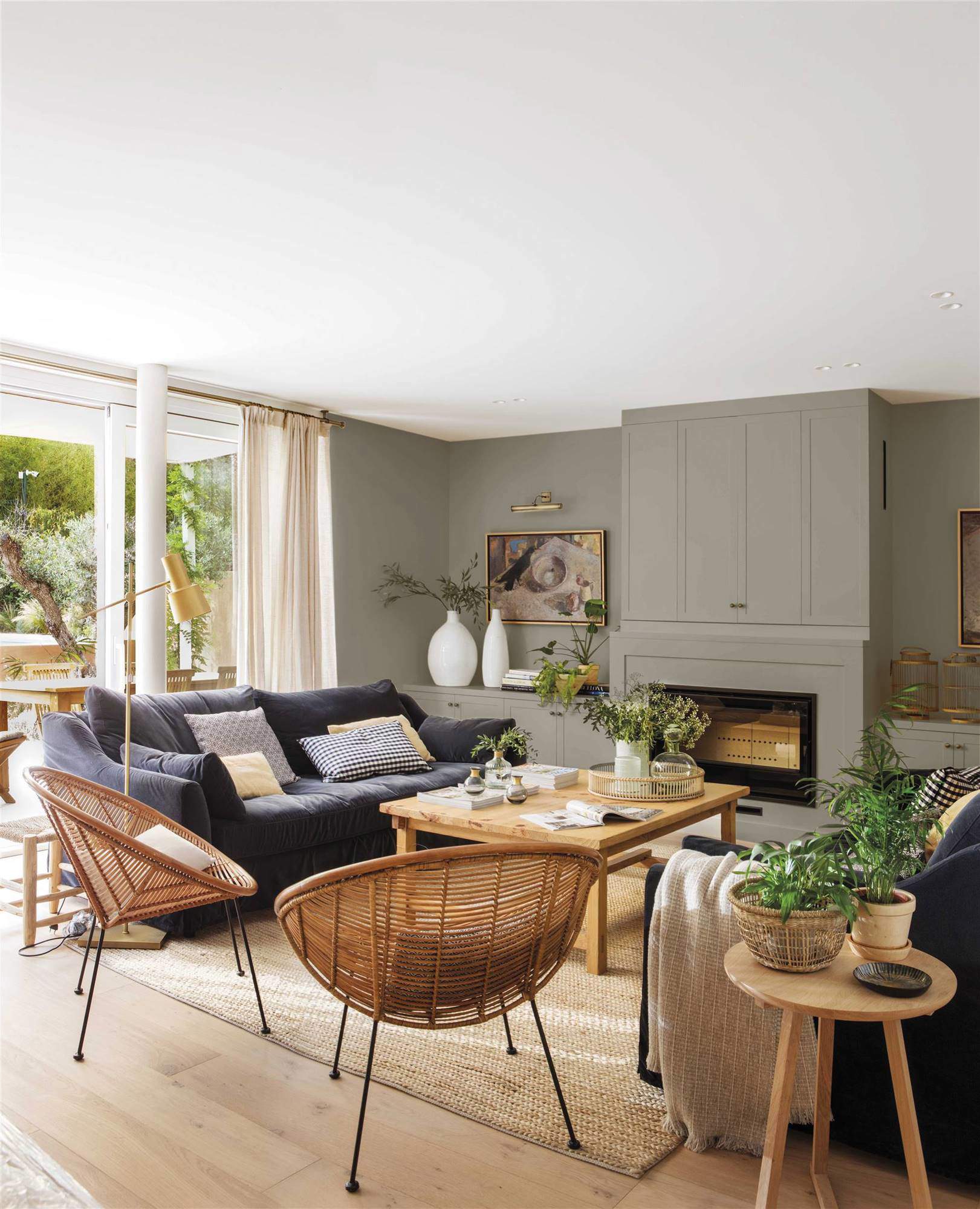 Salón con chimenea y pared principal en gris verdoso, mesa central de madera y sillas en fibras naturales.