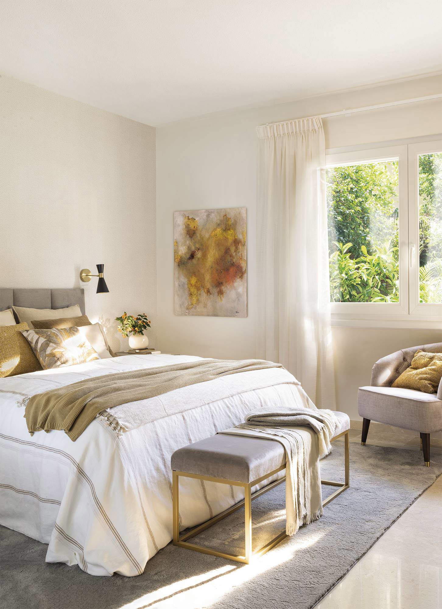 Dormitorio decorado en tonos blancos y dorados, luminoso, con banqueta al pie de la cama.