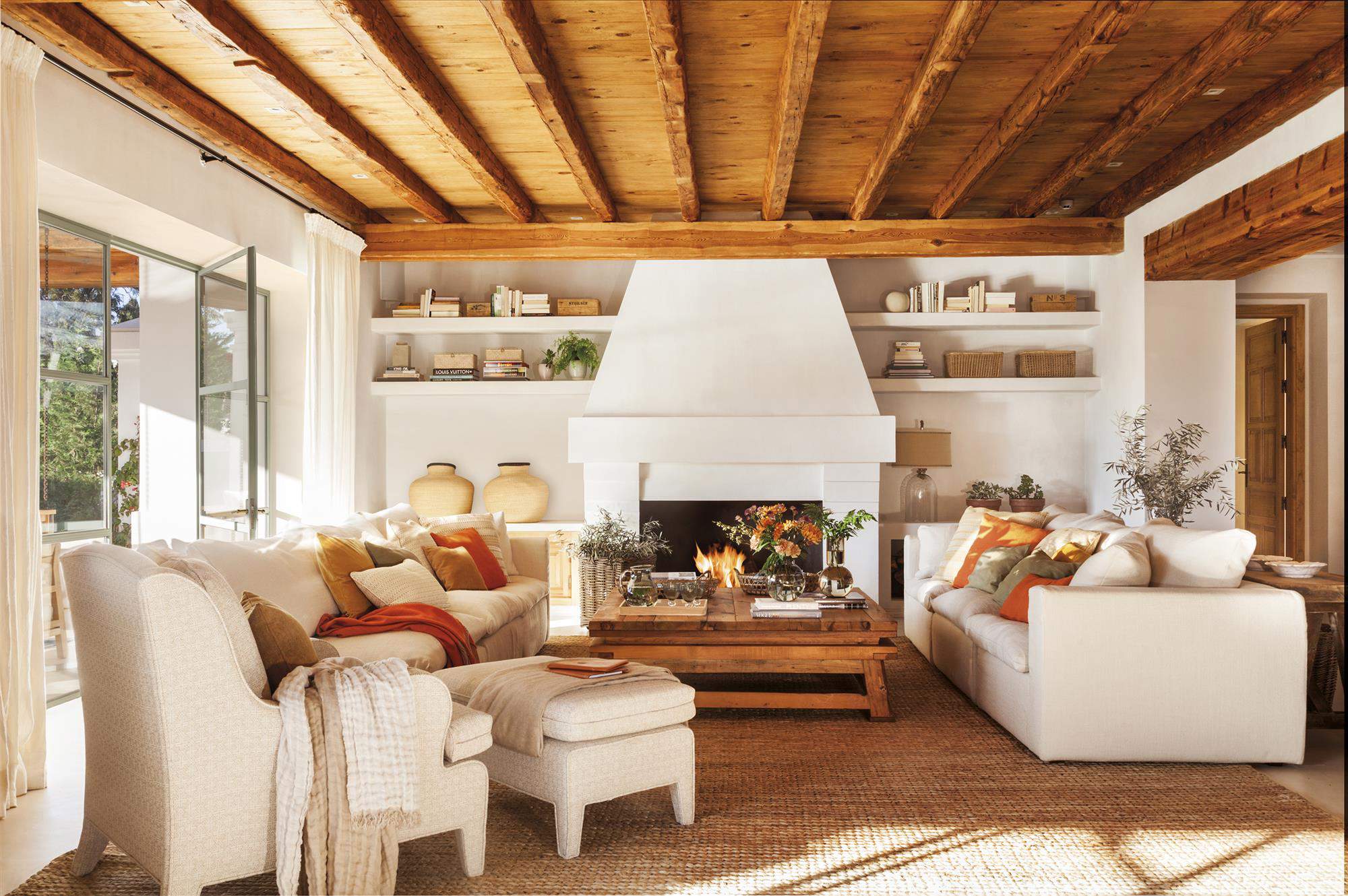 Salón decorado en fibras naturales, chimenea y mobiliario blanco, con vigas de madera natural a la vista.