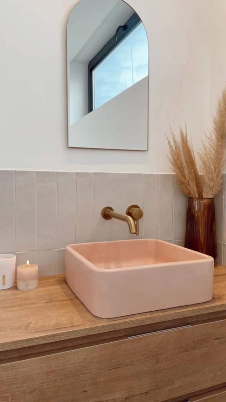 Baño con mueble de madera, lavamanos rosa, grifería dorada y espejo. 