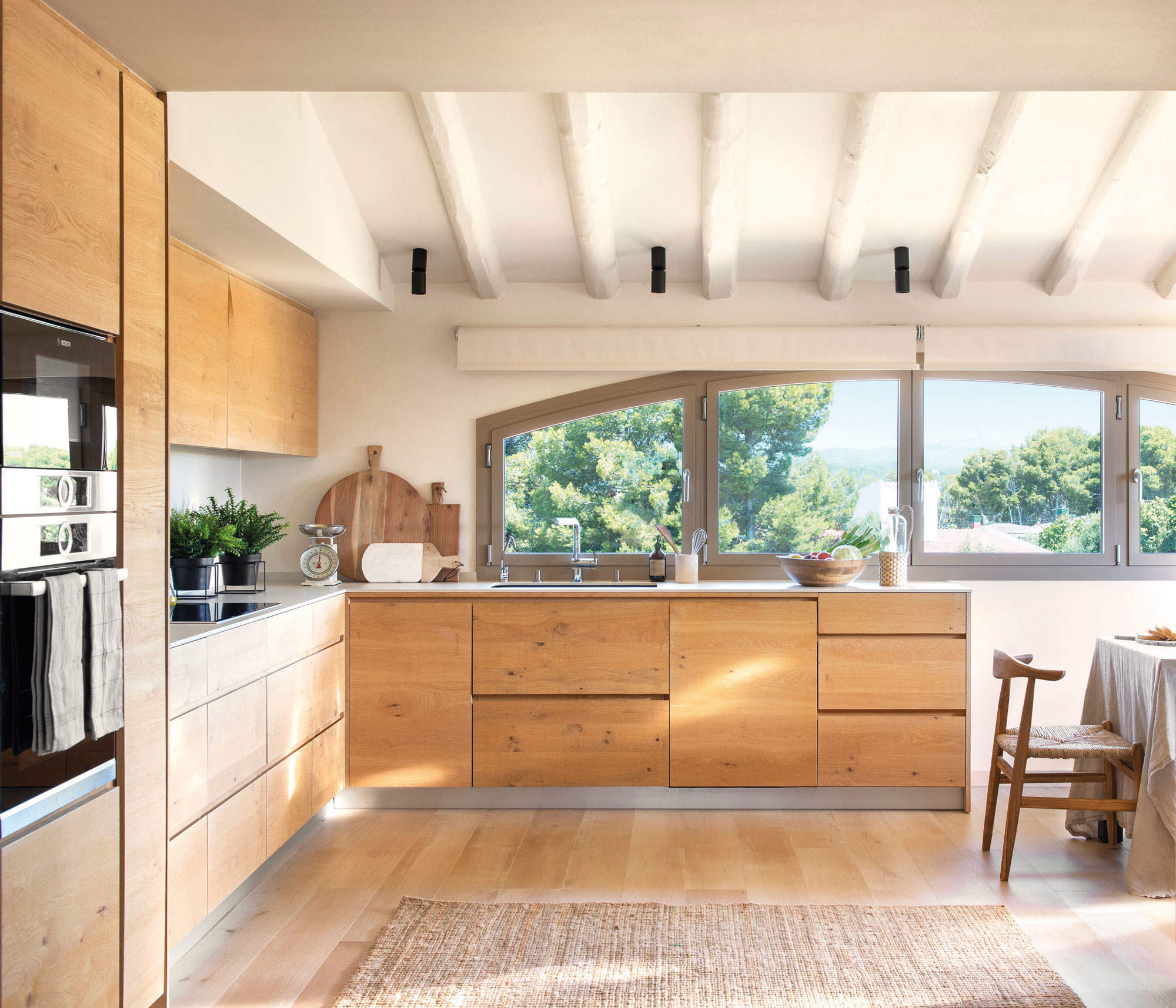 Cocina con ventana de semiarco, muebles de madera natural y vigas blancas a la vista.