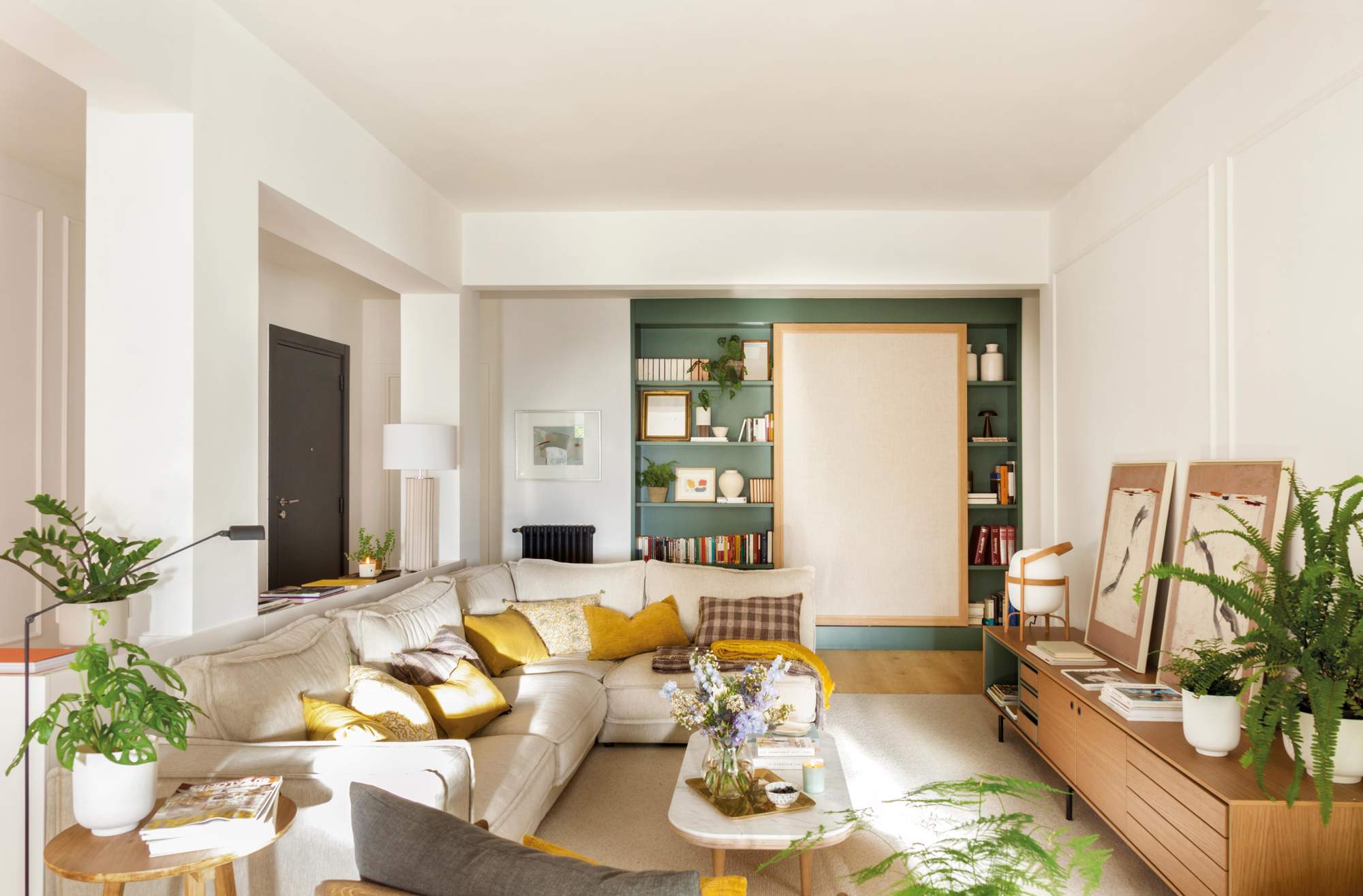 Salón con sofá esquinero, estantería pintada de verde, mueble bajo, cuadros y plantas.