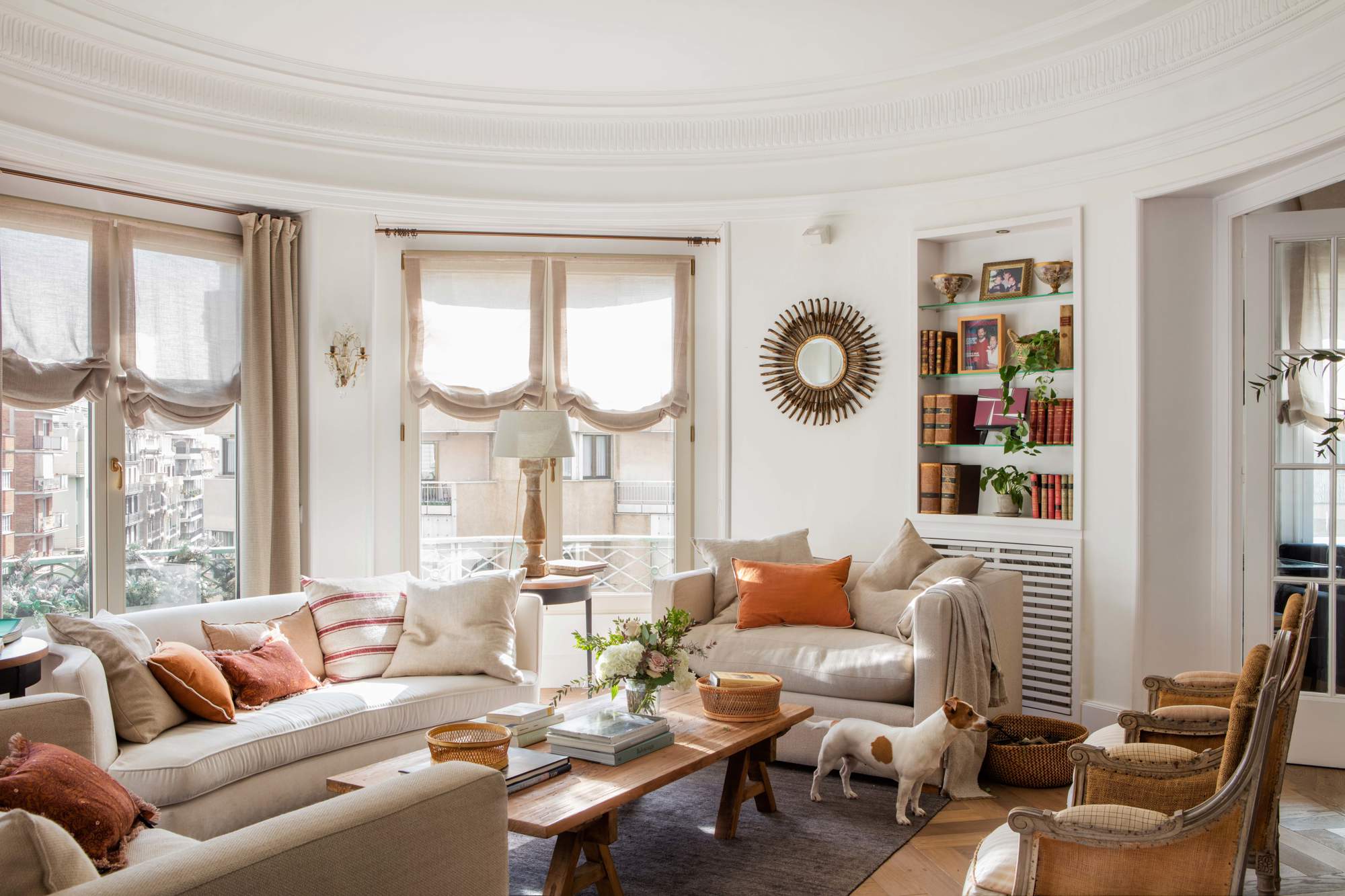 Salón clásico con sofás blancos, mesa de madera, butacas, estantería, espejo sol, estores y perro.