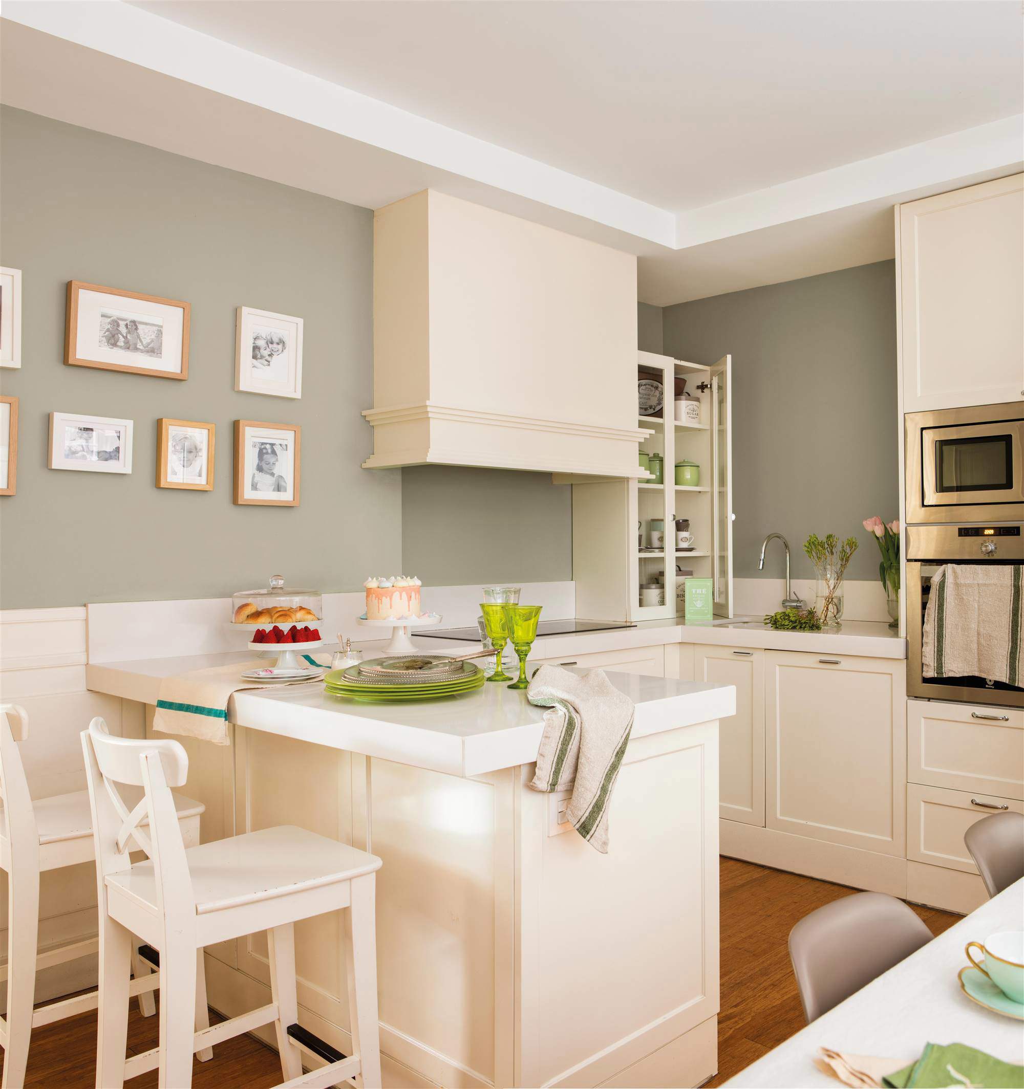Cocina blanca con península a modo de barra y comedor, pared pintada en verde grisáceo, campana y composición de fotos familiares.