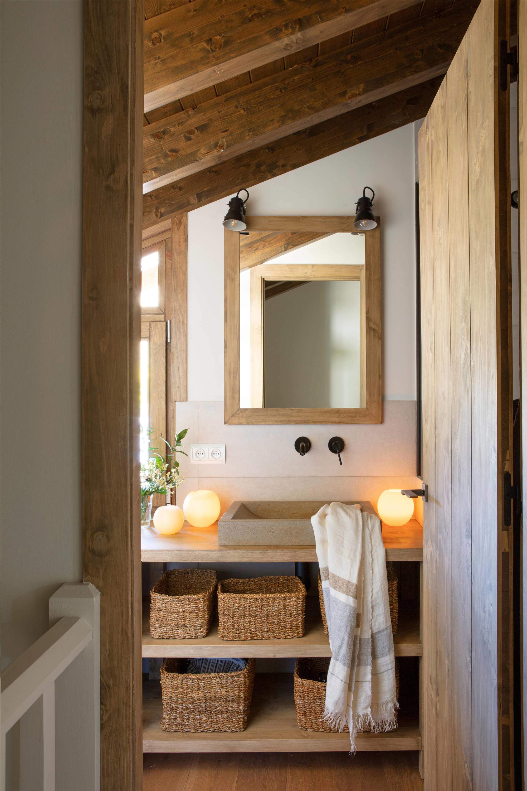 Baño con mueble bajolavabo abierto y de madera, grifería negra, espejo de madera, apliques negros, cestas de fibra y velas redondas.