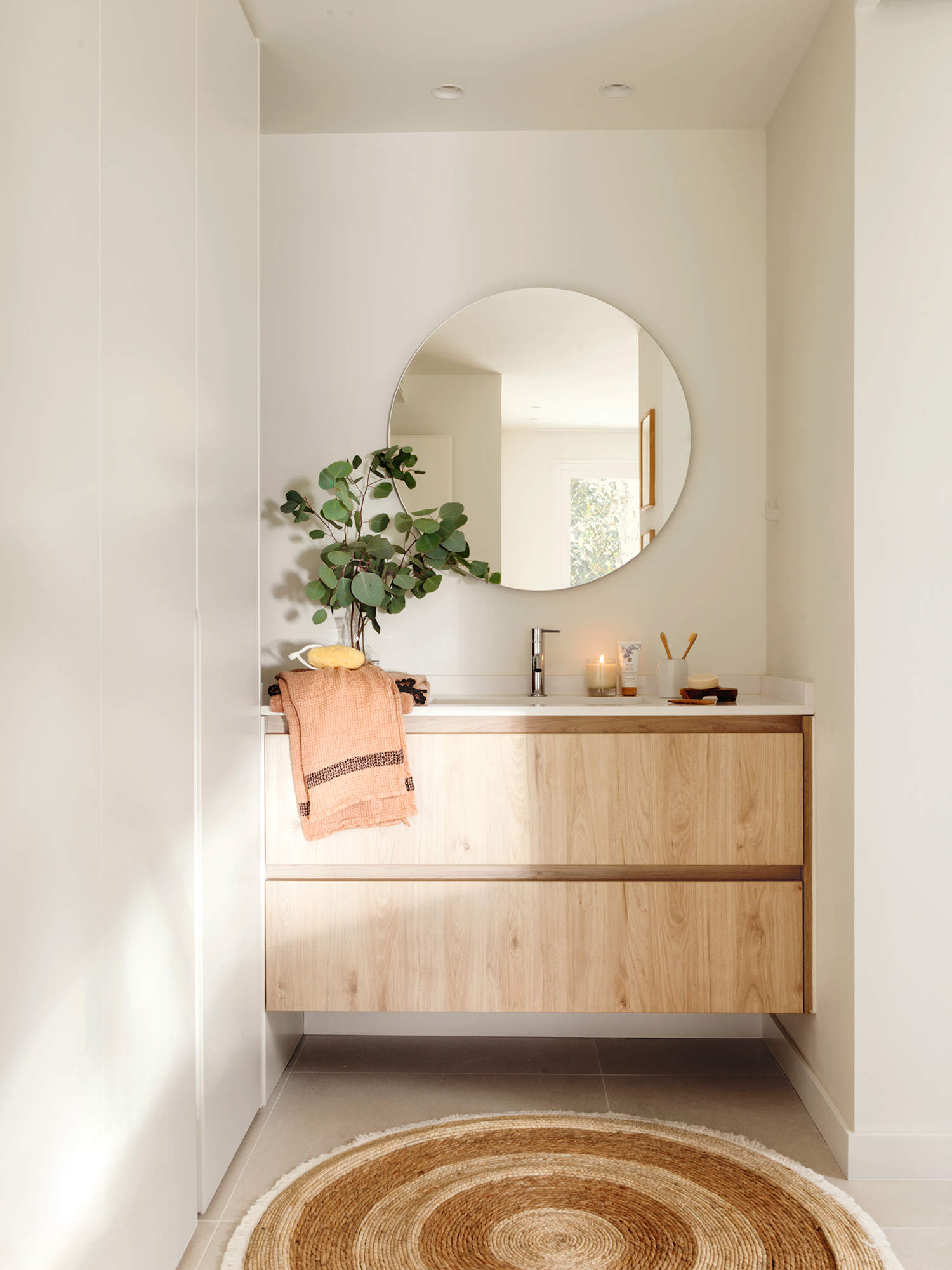 Baño pequeño con mueble realizado a medida, espejo redondo, plantas y alfombra de fibra.