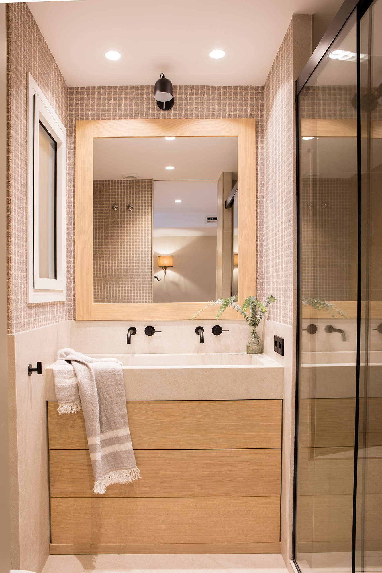 Mueble de baño encastrado entre paredes, papel pintado de cuadros marrones y mampara acristalada ahumada. 