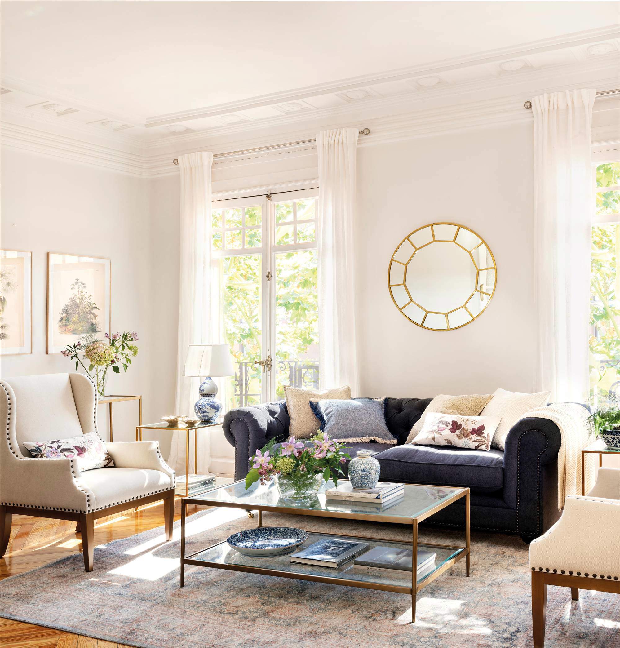 Salón con sofá azul marino, cojines claros, butacas en beige, espejo dorado, mesa de centro en cristal y metal, alfombra y cuadros